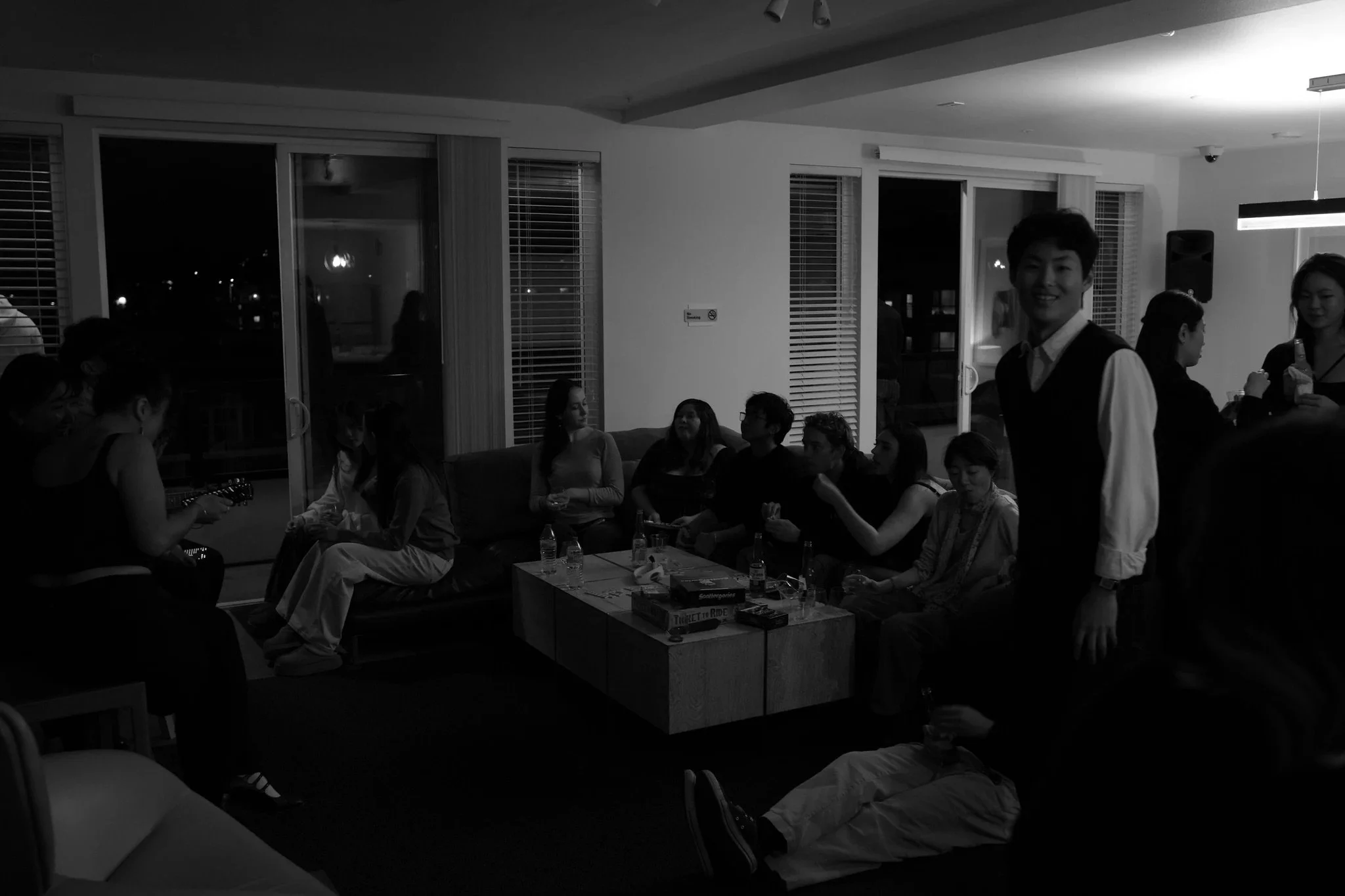 People socializing at an indoor gathering, some sitting on couches and others standing, in a room with large windows and a modern decor.