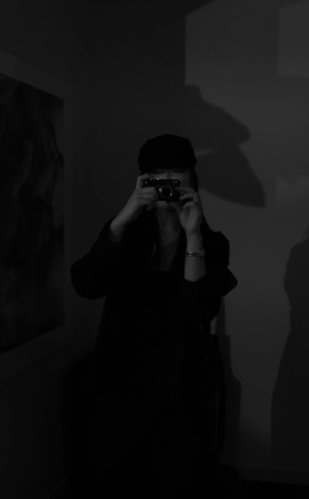 A person takes a mirror selfie with a camera, wearing a hat and a watch, in a dimly lit room.
