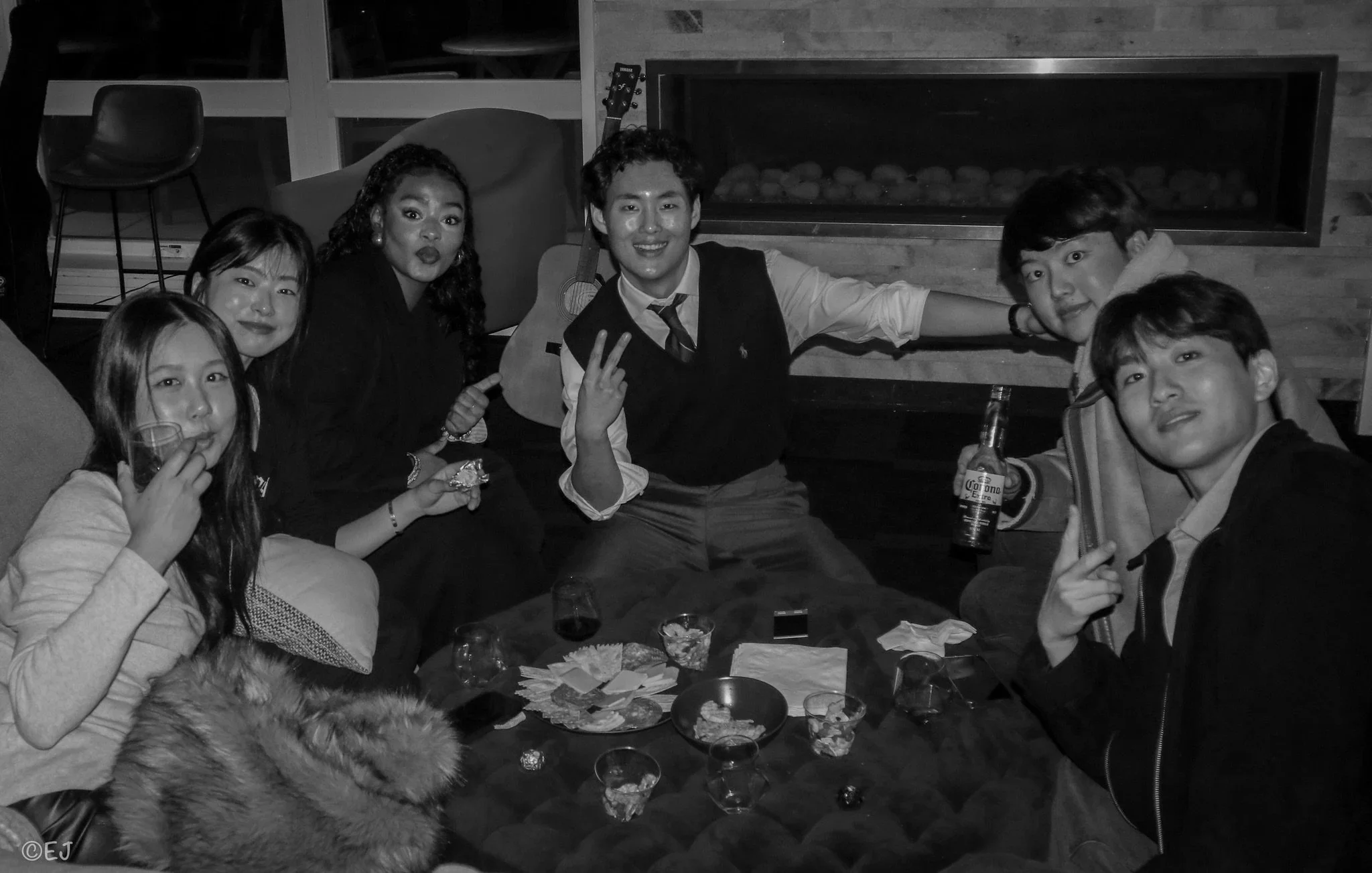 Group of six young adults at a party, sitting around a table with drinks and snacks, some making peace signs, smiling, and enjoying the night in a cozy indoor setting.