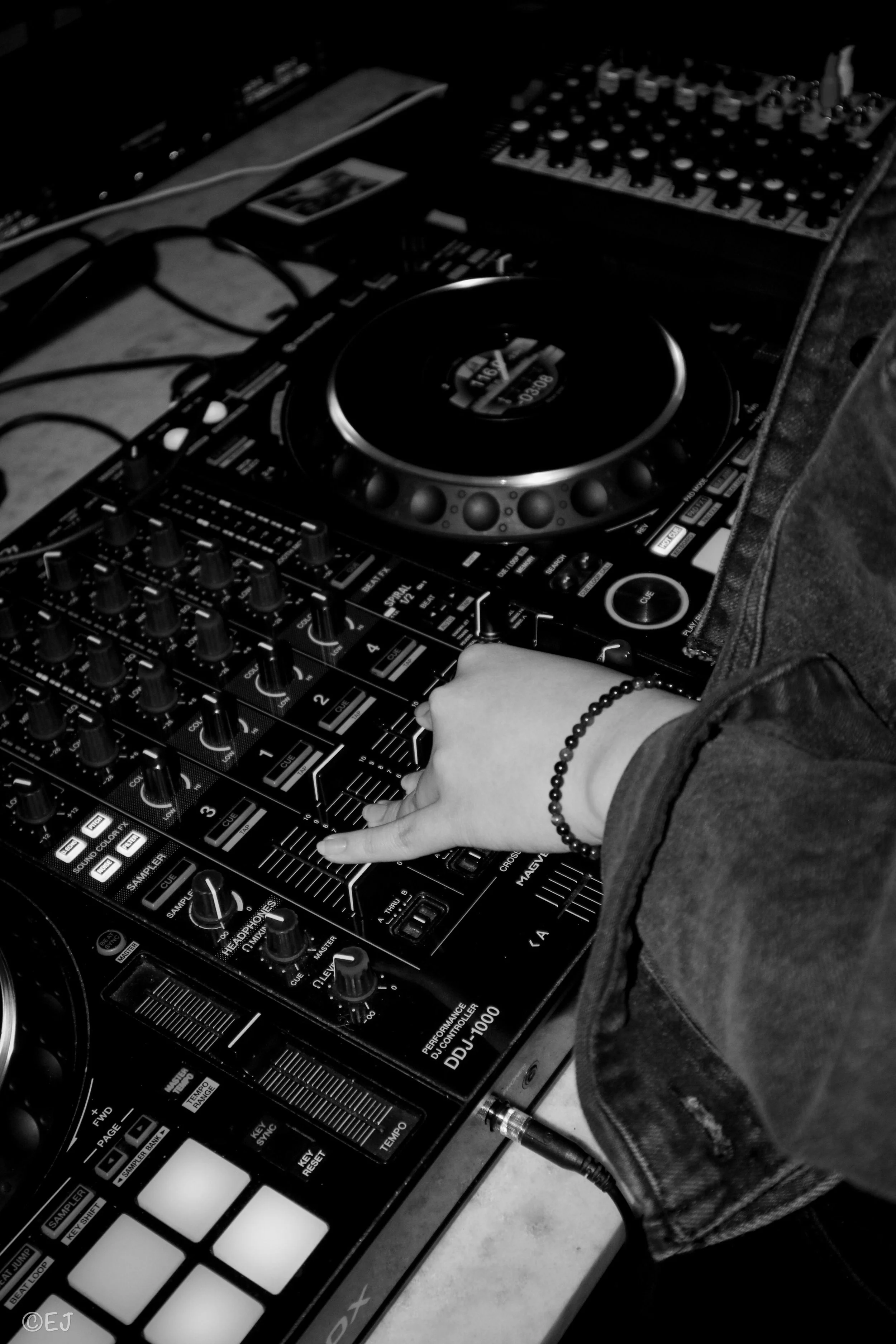 A person's hand is on a DJ controller with various knobs, buttons, and a jog wheel, surrounded by audio equipment and cables.
