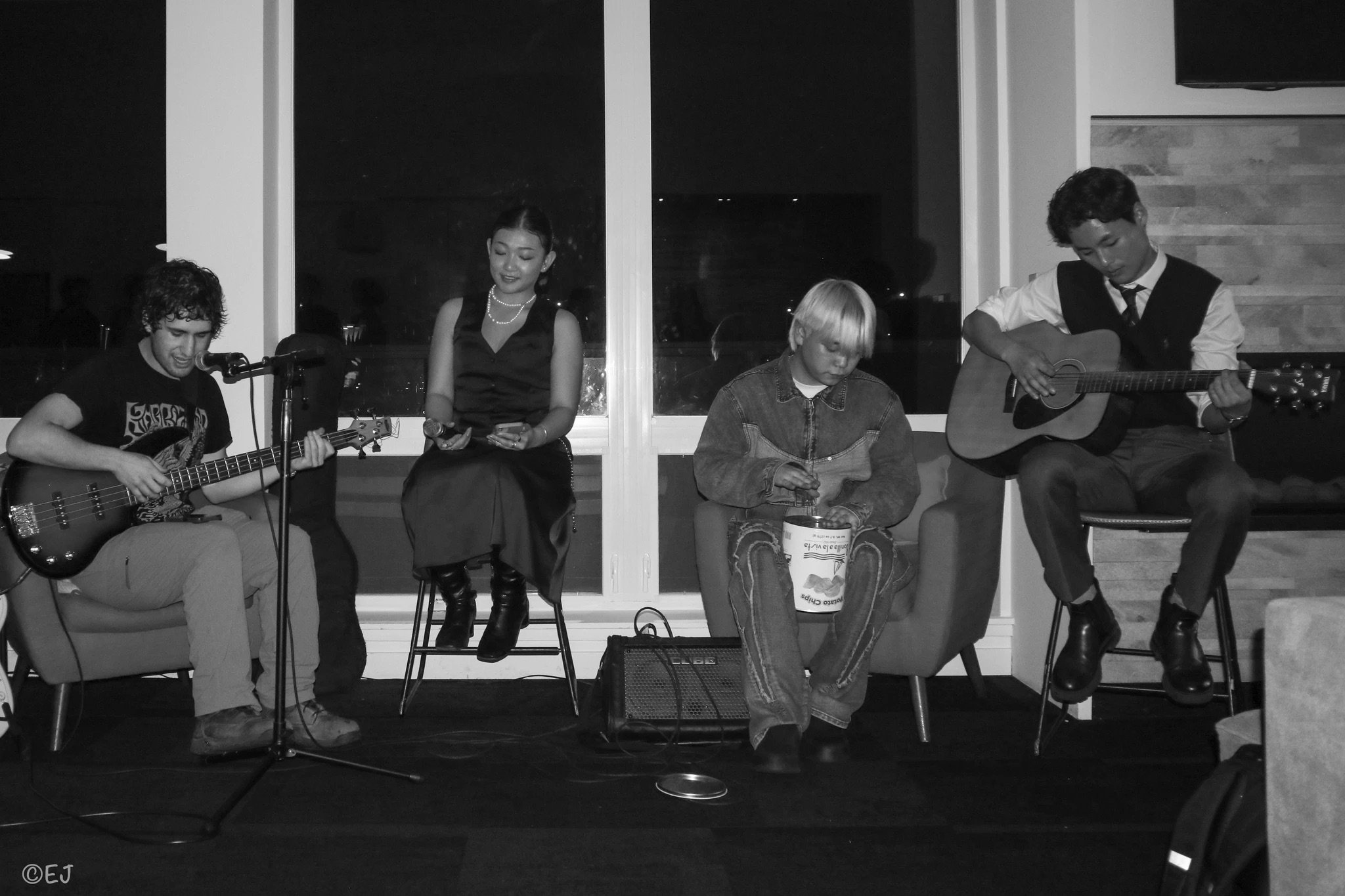 A group of four young people performing music and singing indoors at night, with two playing guitars, one singing, and one sitting with a drum. All are seated in front of large windows.