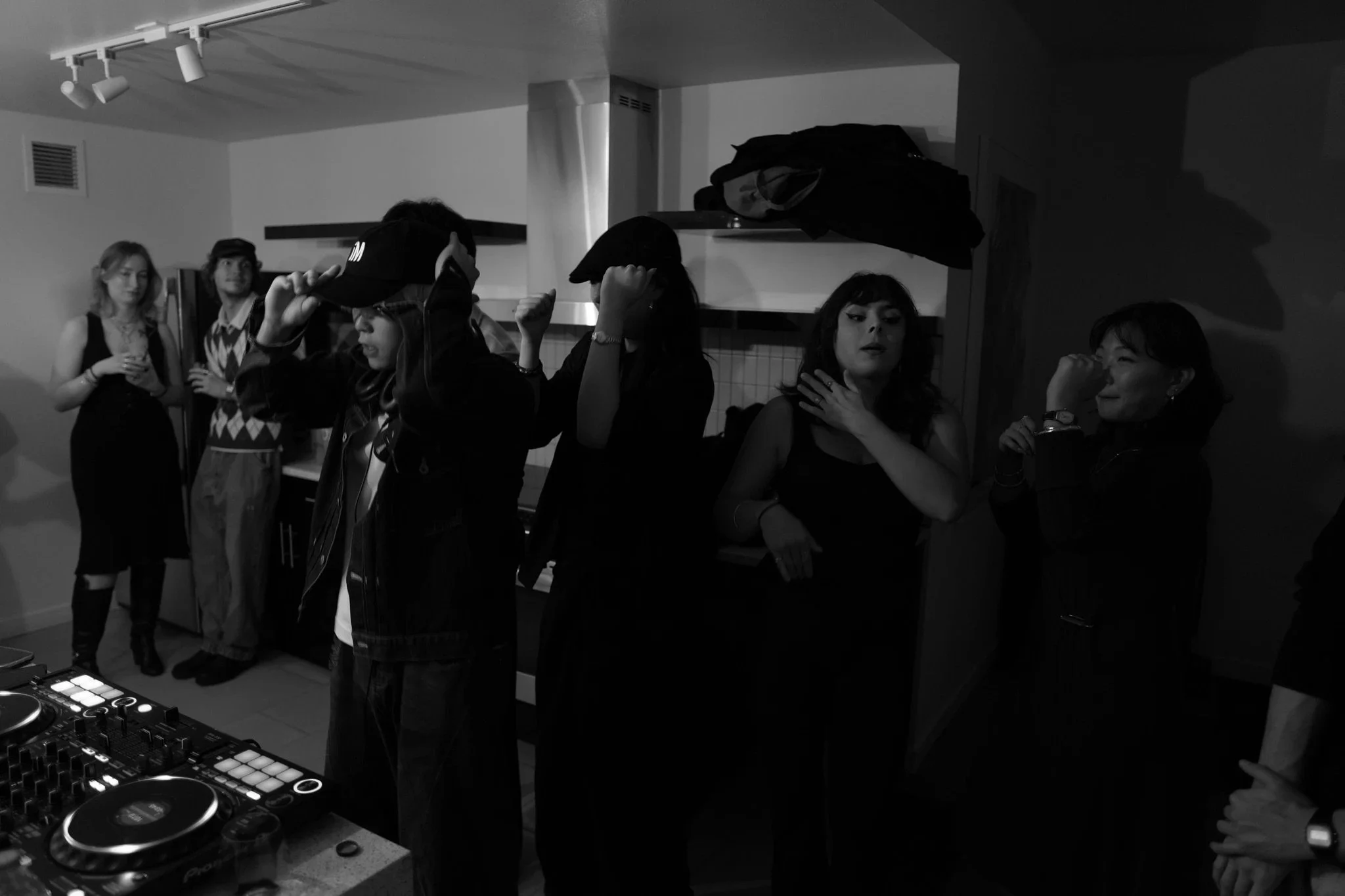 A group of people at a gathering, some with drinks, some adjusting their hats, standing in front of a countertop with DJ equipment.