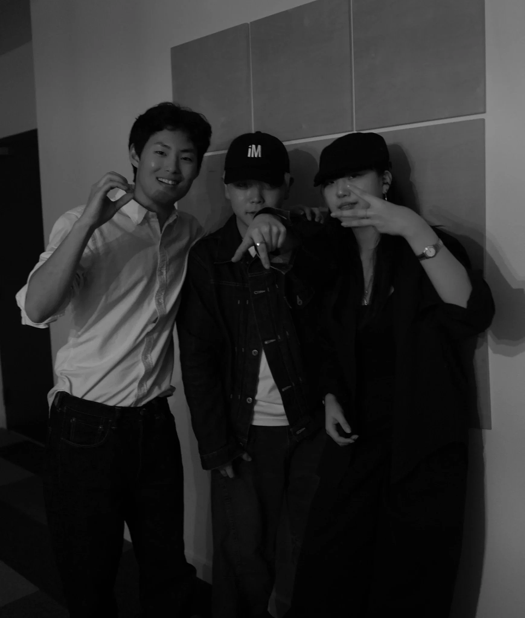 Three young men posing together indoors against a wall, with one wearing a cap labeled 'iM', and making hand gestures.