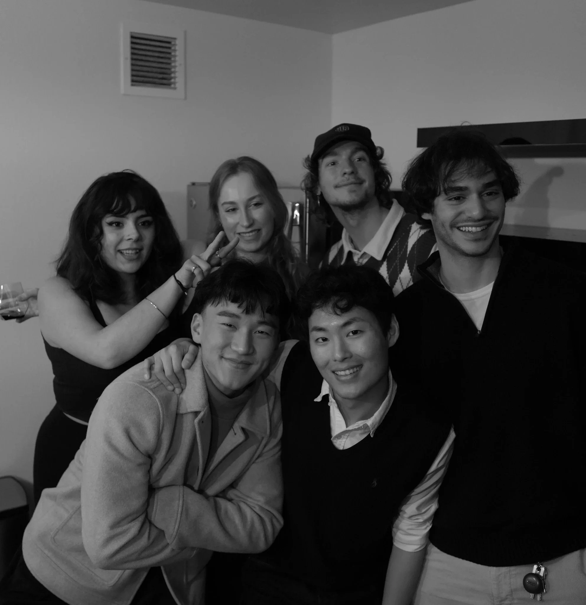 Group of five friends smiling and posing for a photo indoors. Four are clearly visible, three men and one woman, all smiling. The woman has wavy hair and is making a peace sign. The men have curly and straight hair. Some are wearing sweaters, and the