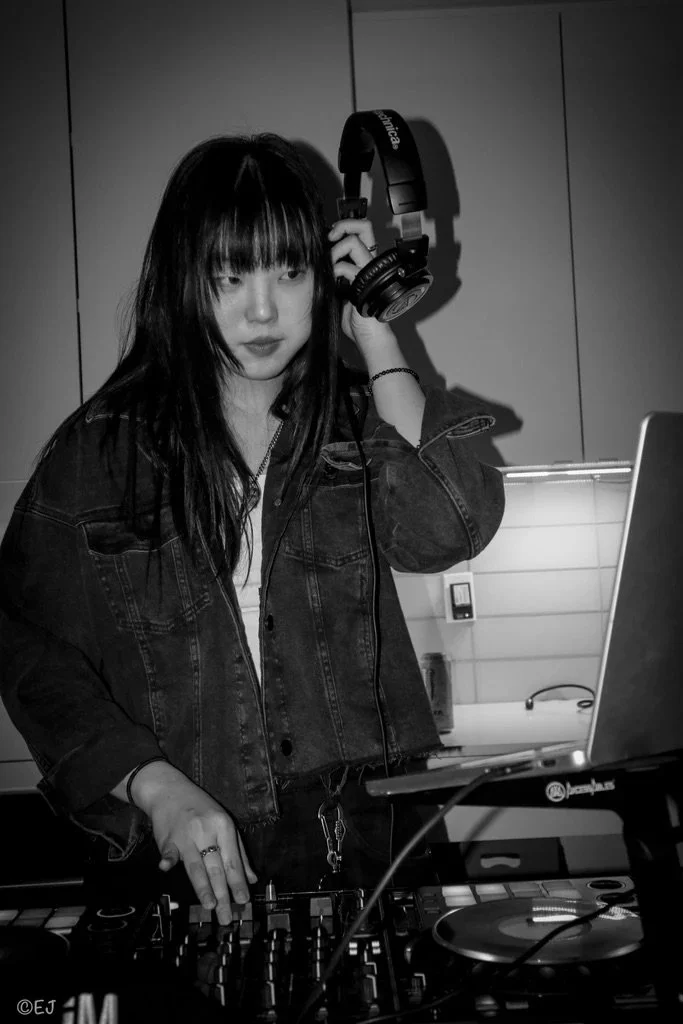 A woman with dark hair and bangs wearing a denim jacket, holding headphones in her right hand, standing at a DJ setup with a laptop and mixer, in an indoor space with cabinets in the background.