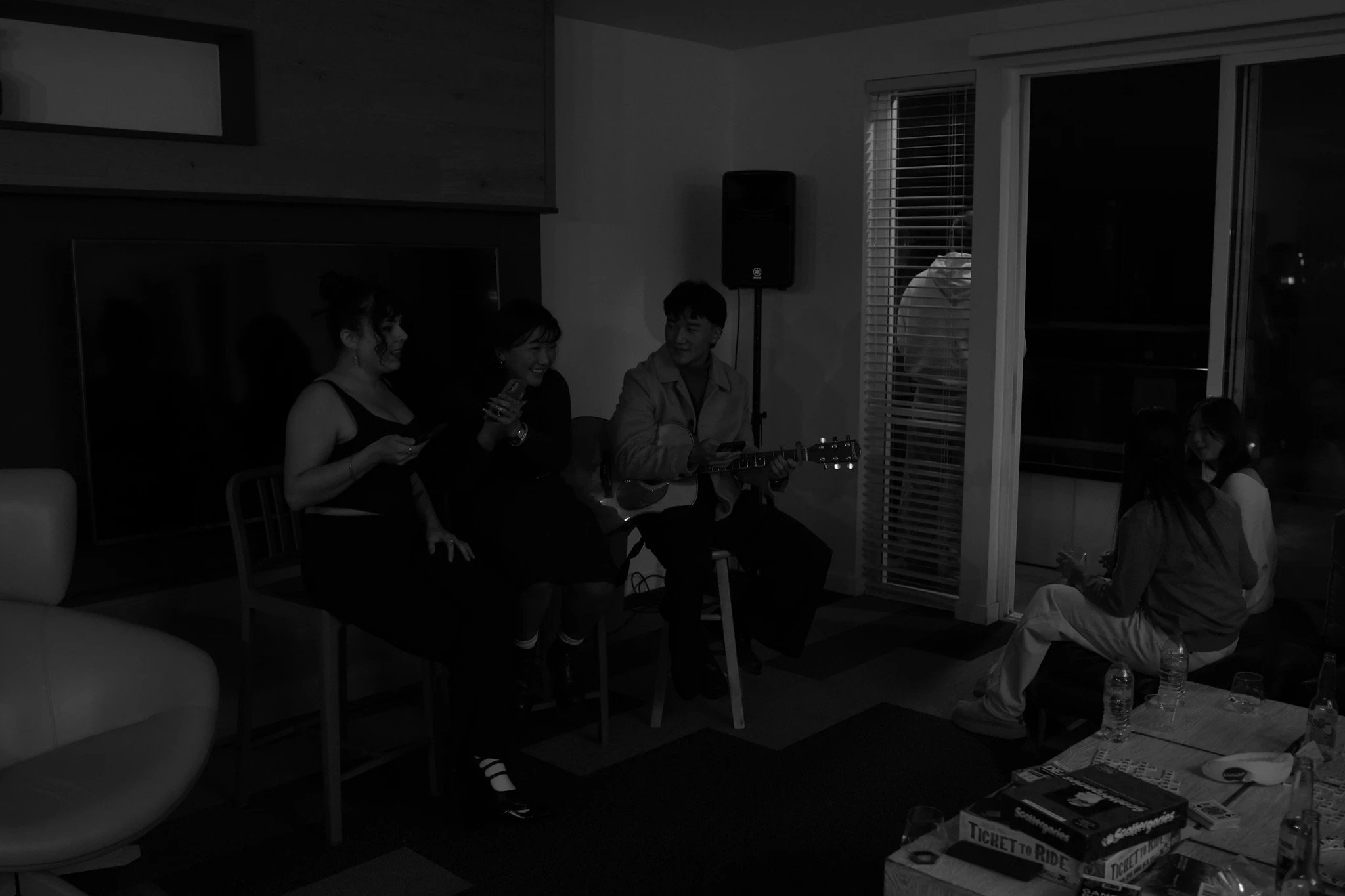 A group of people sitting in a living room, some holding musical instruments and others chatting, with a table of board games and bottled water on it.