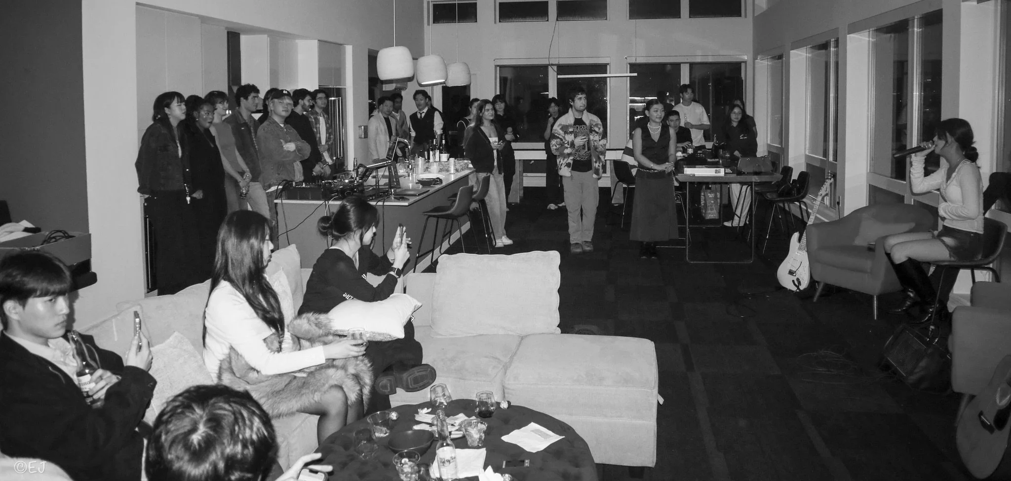 A group of people gathered indoors, some sitting on couches and chairs, others standing near a table with drinks and snacks, while a woman on the right is singing into a microphone, accompanied by a guitar on a stand. The setting appears to be a soci