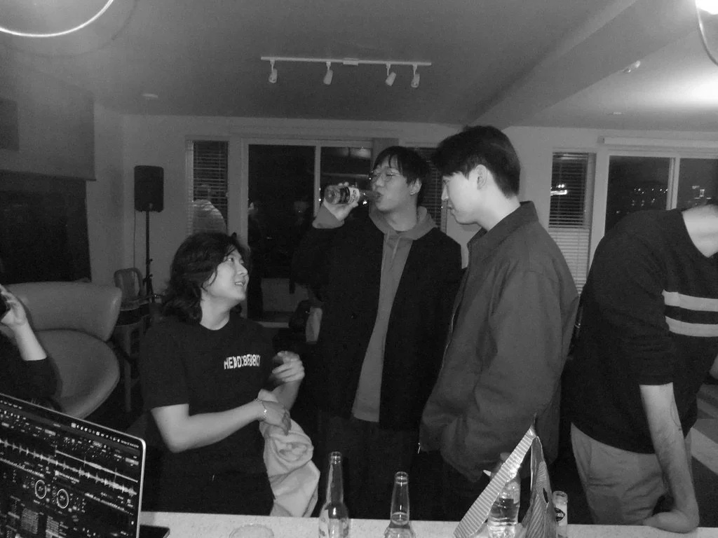 Group of young people socializing indoors, with three people standing and one sitting, engaged in conversation and drinking beer, in a living room setting with large windows and modern decor.