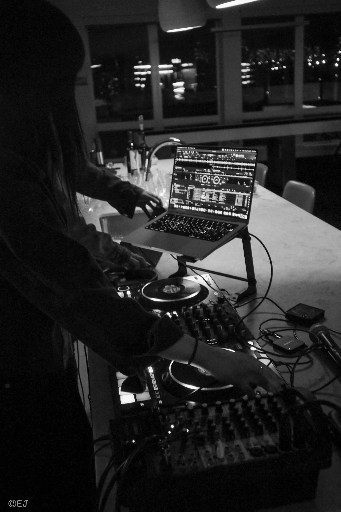 A person DJing at a table with a laptop and DJ equipment in a dimly lit room, with city lights visible through large windows in the background.