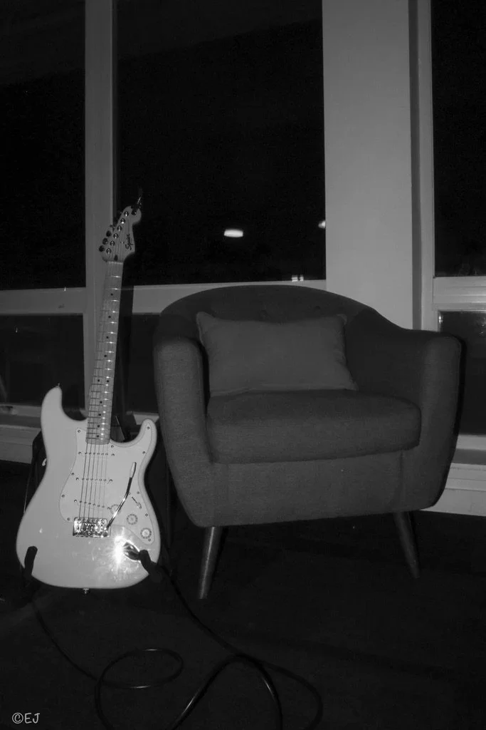 An electric guitar and a cozy armchair on a dark indoor floor next to a window.