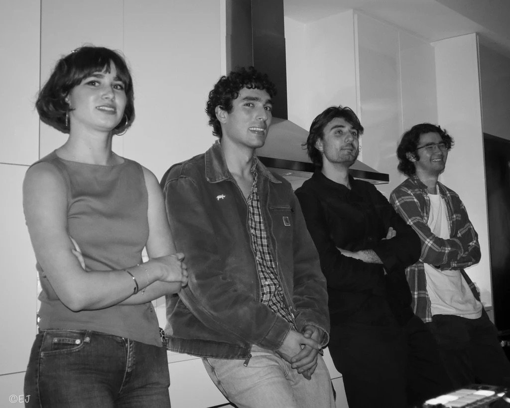Four young adults standing in a row, smiling and leaning against a kitchen counter, in a modern kitchen with white cabinets.