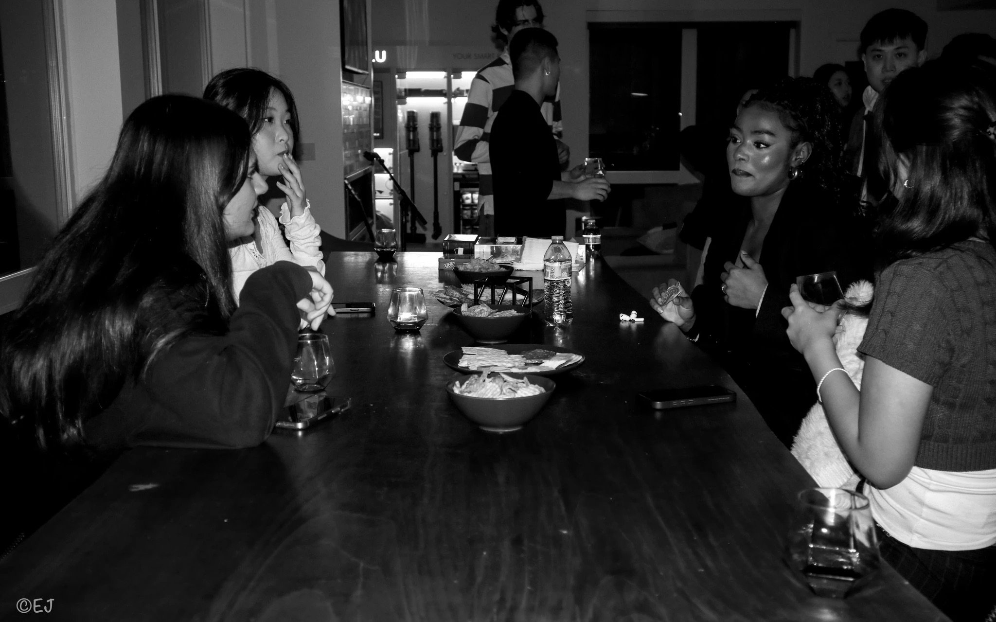 A group of people sitting at a dining table in a restaurant, engaging in conversation while enjoying food and drinks, with a bartender or waiter in the background.