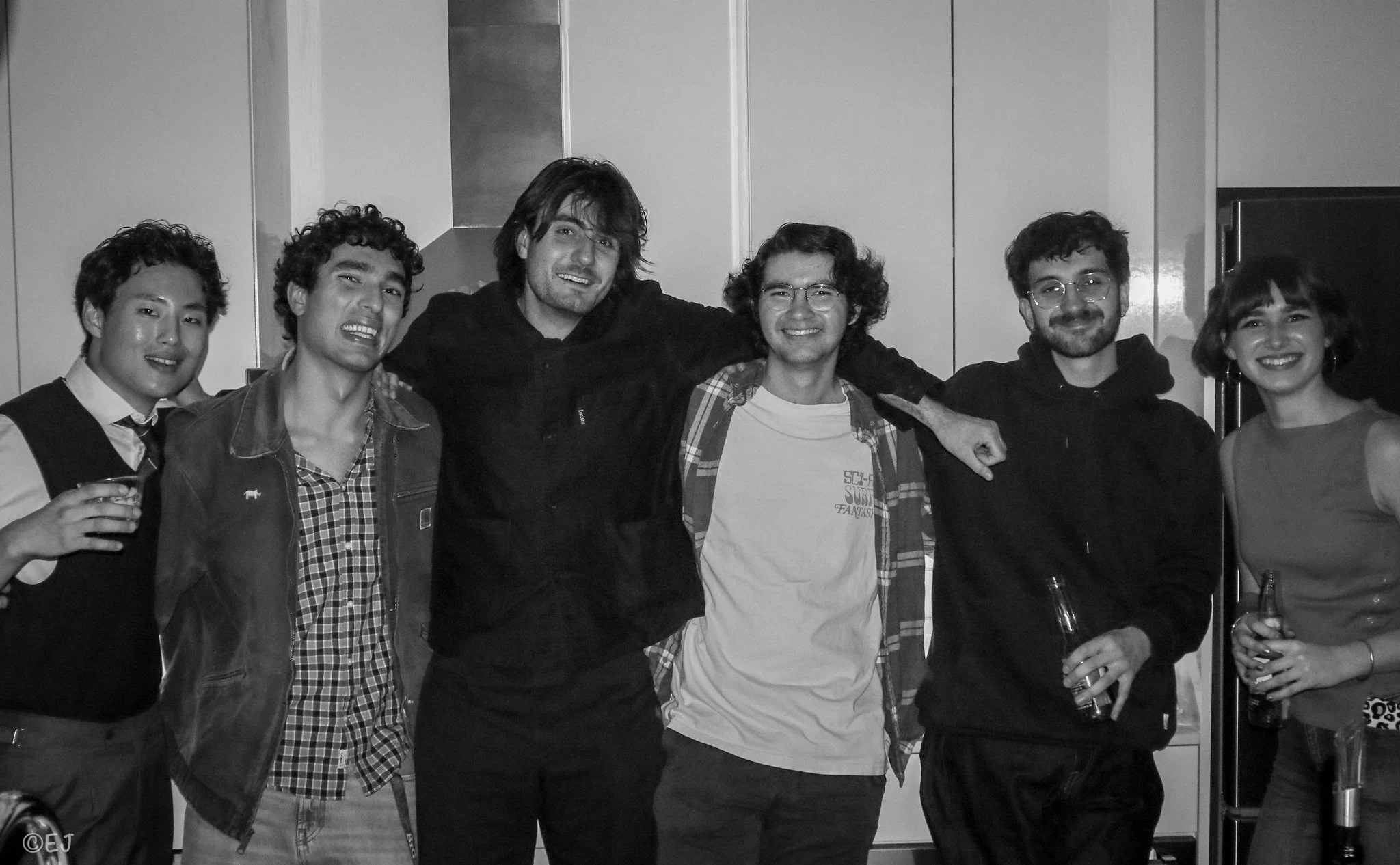 Group of six young adults smiling, standing close together, arms around each other in a casual indoor setting.