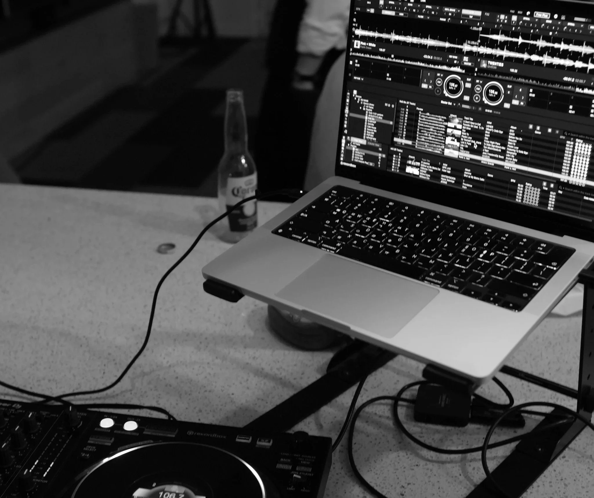 A laptop with DJ software on the screen, a partially visible DJ controller in the foreground, a clear glass bottle of Corona beer, and a coin on a gray surface.