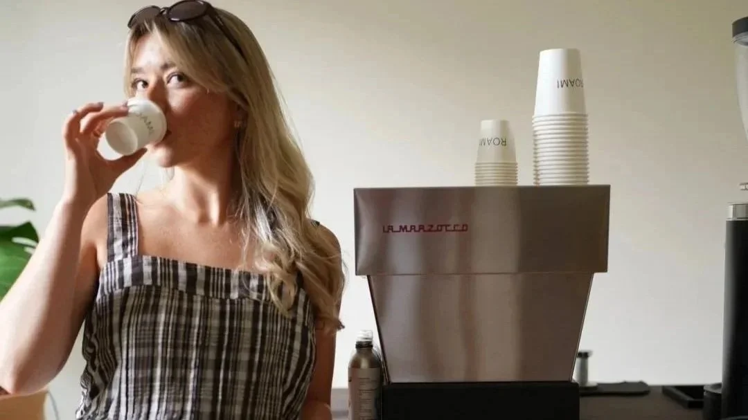 Woman drinking espresso beside a La Marzocco coffee machine and stacked cups on the coffee cart