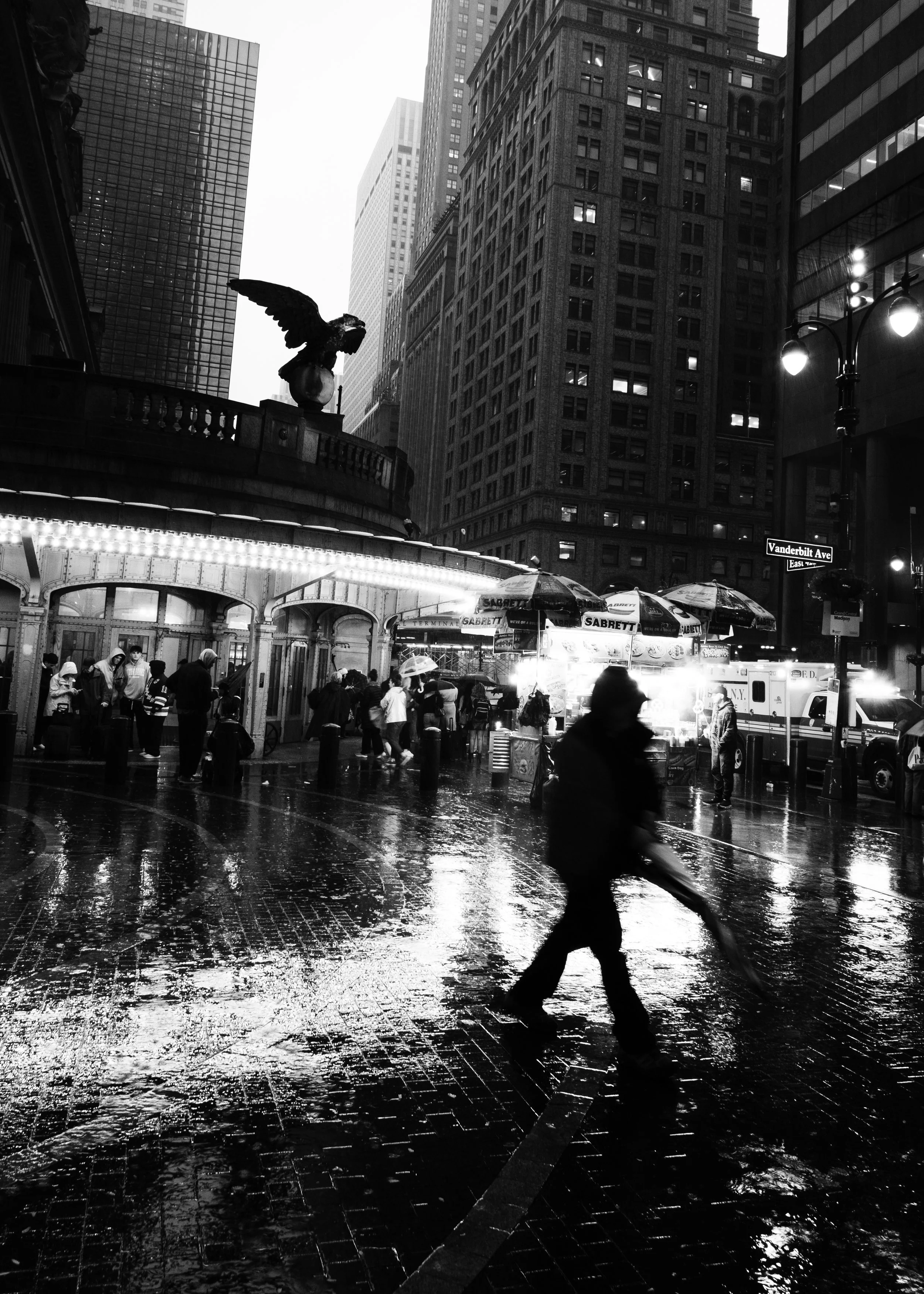 A busy city street scene during rainy weather at night. People are walking with umbrellas, and a person is crossing the street on a scooter. Tall buildings and a street sign indicating Vanderbilt Ave are visible in the background.