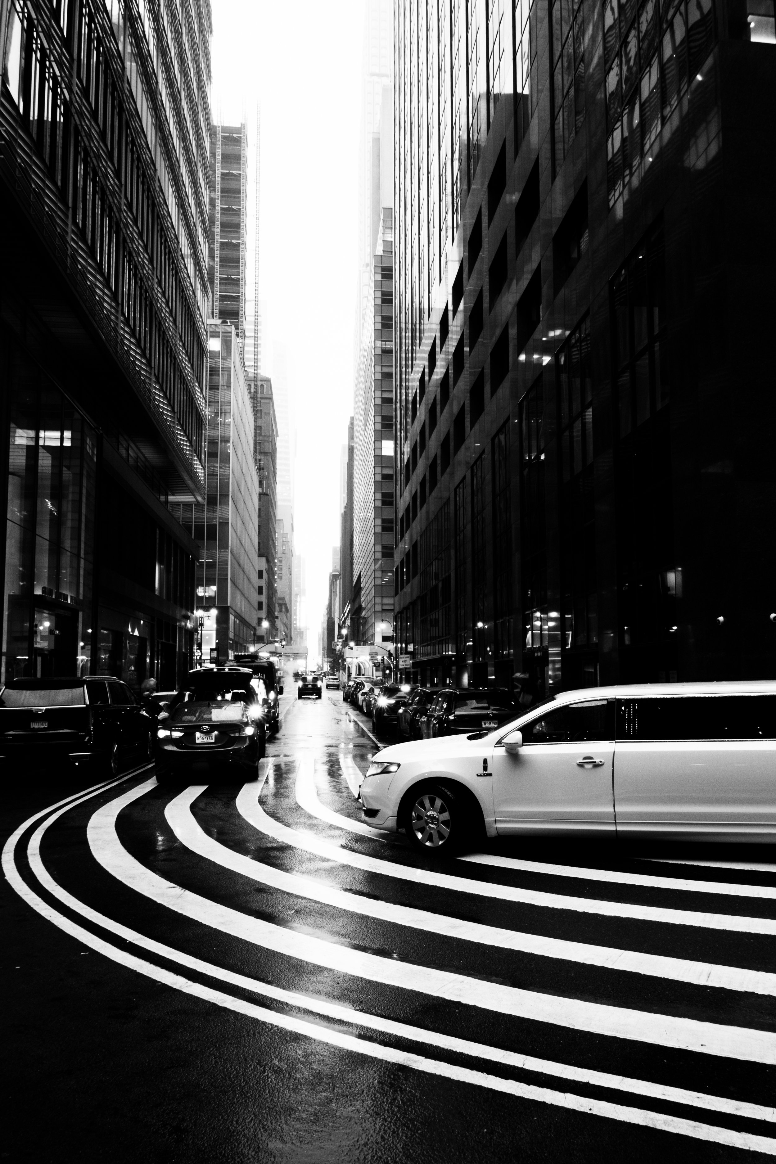Black and white city street scene with tall glass buildings on both sides, cars parked along the street, and curved line markings on the wet road surface.
