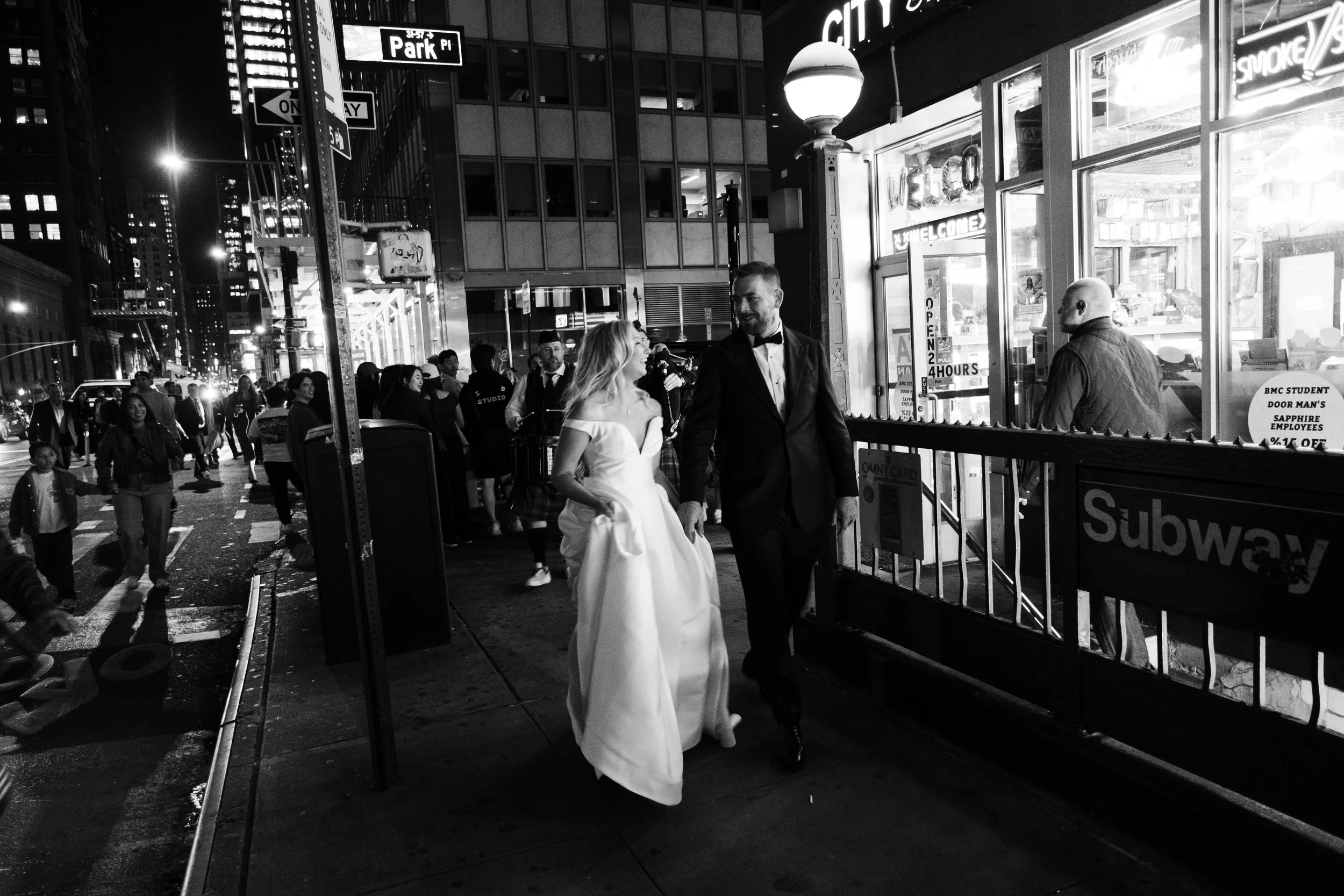 A bride and groom in evening attire walking on a city sidewalk, with a crowd behind them and illuminated storefronts around. The bride wears a white dress and the groom a black tuxedo. The scene is in black and white.