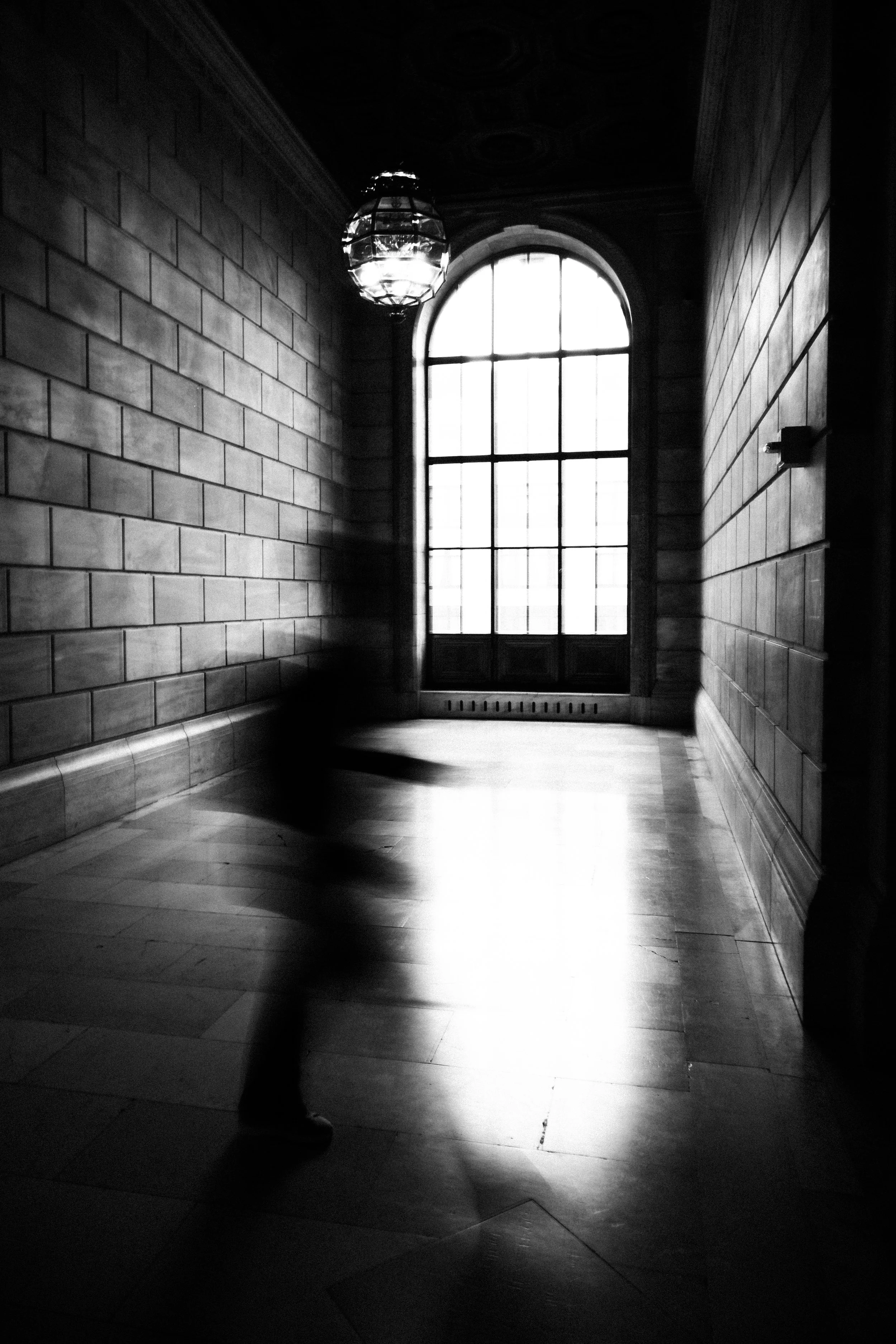 Black and white photo of a hallway with brick walls, a large arched window, and a hanging ceiling light fixture. A shadow of a person is cast on the floor.