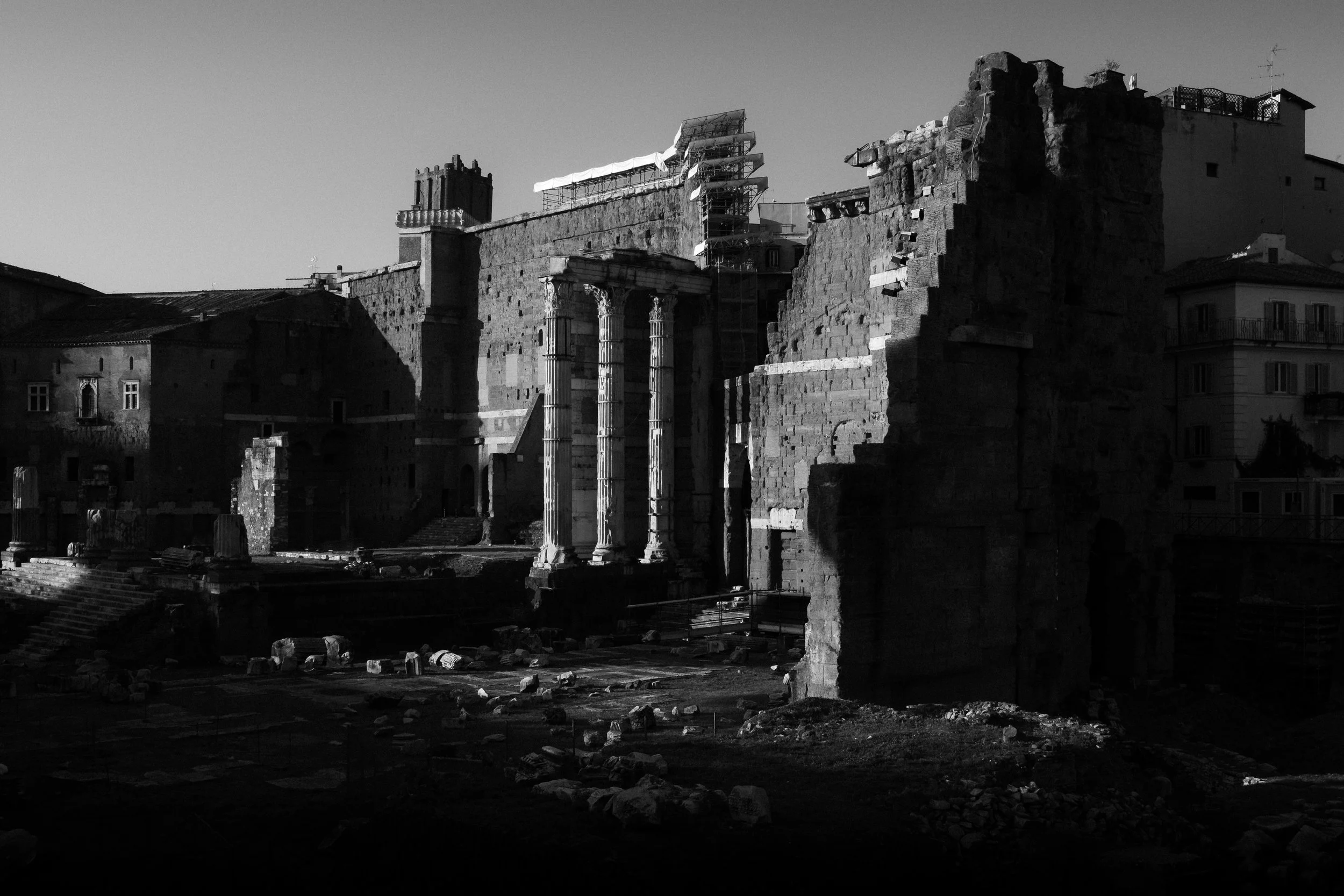 Ancient ruins with partially standing columns and crumbling stone walls, some buildings and scaffolding in the background, under a clear sky.