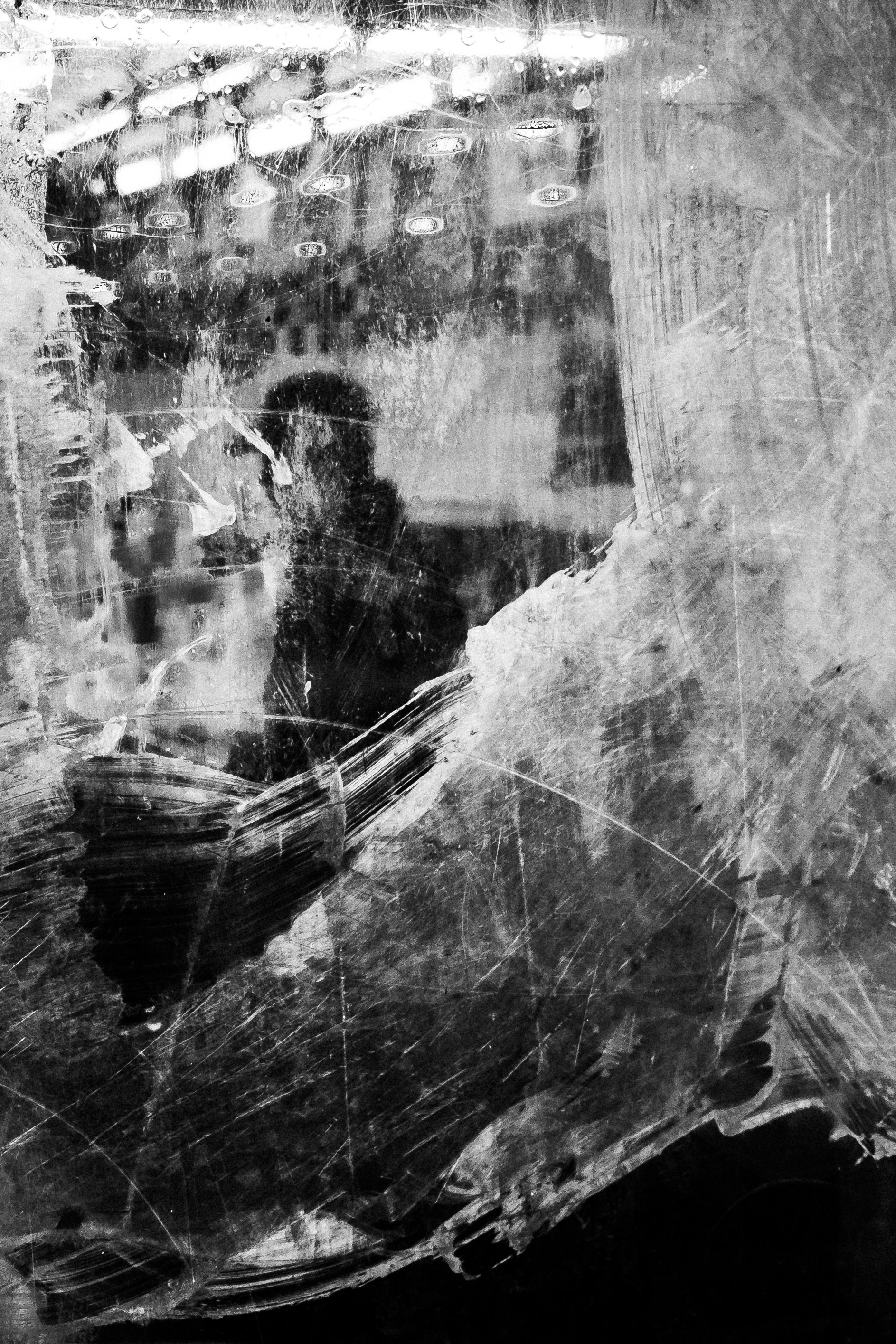A black and white photo of a person taking a selfie in a dirty and scratched mirror. The person's face is blurry and partially visible, with a hat or cap on their head. The background shows various reflections and smudges.