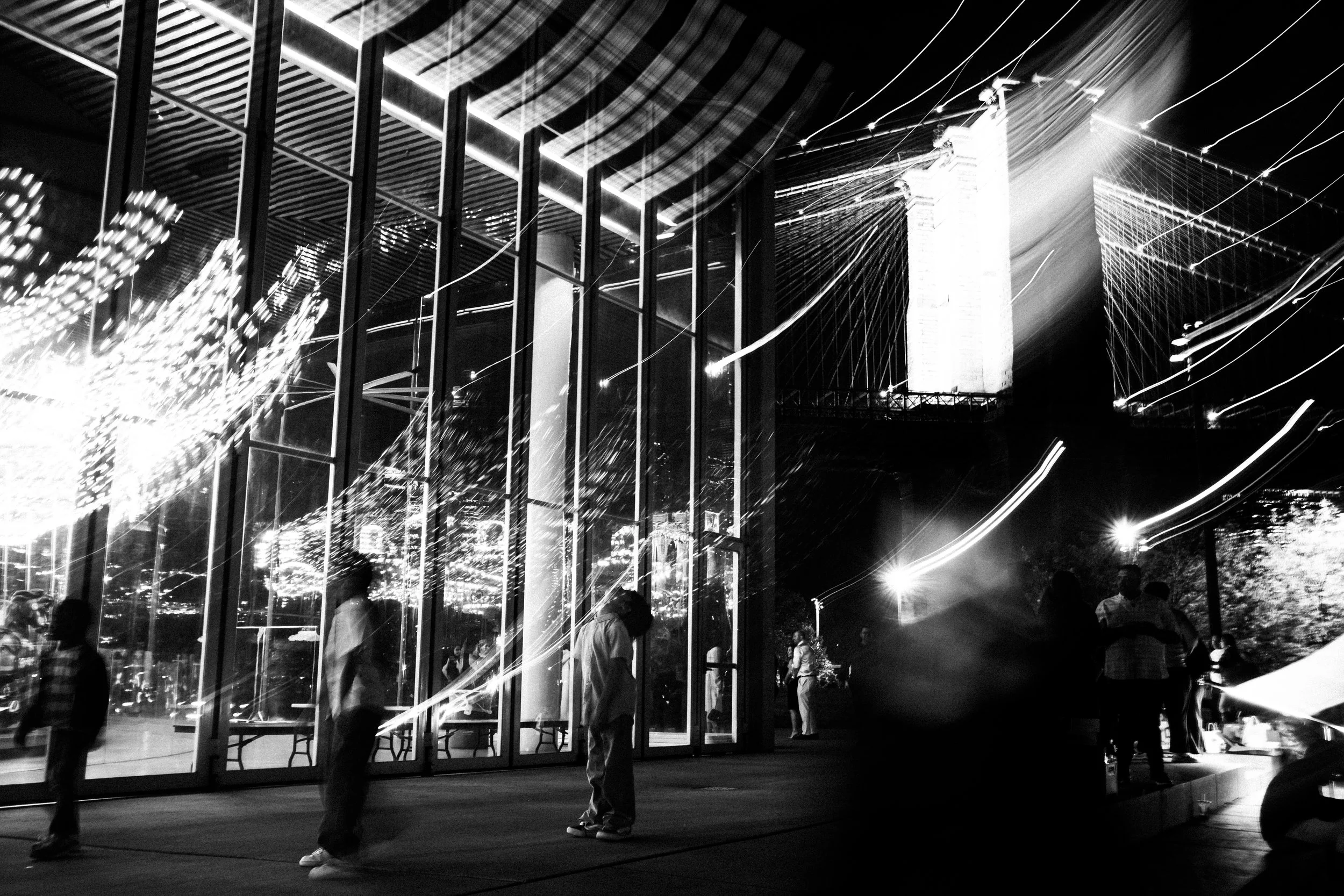 Nighttime scene of modern glass buildings with lights, with people walking outside and streaks of light from long exposure.
