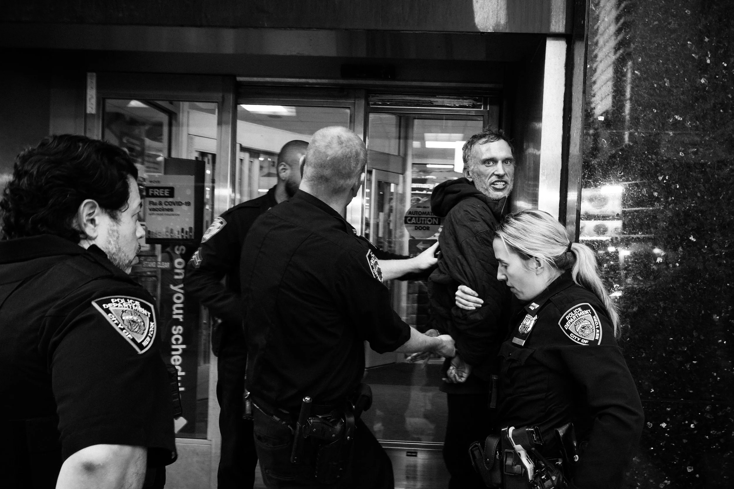 Group of police officers, one man actively speaking or yelling, inside a building with glass doors and windows, some officers are looking down or to the side.