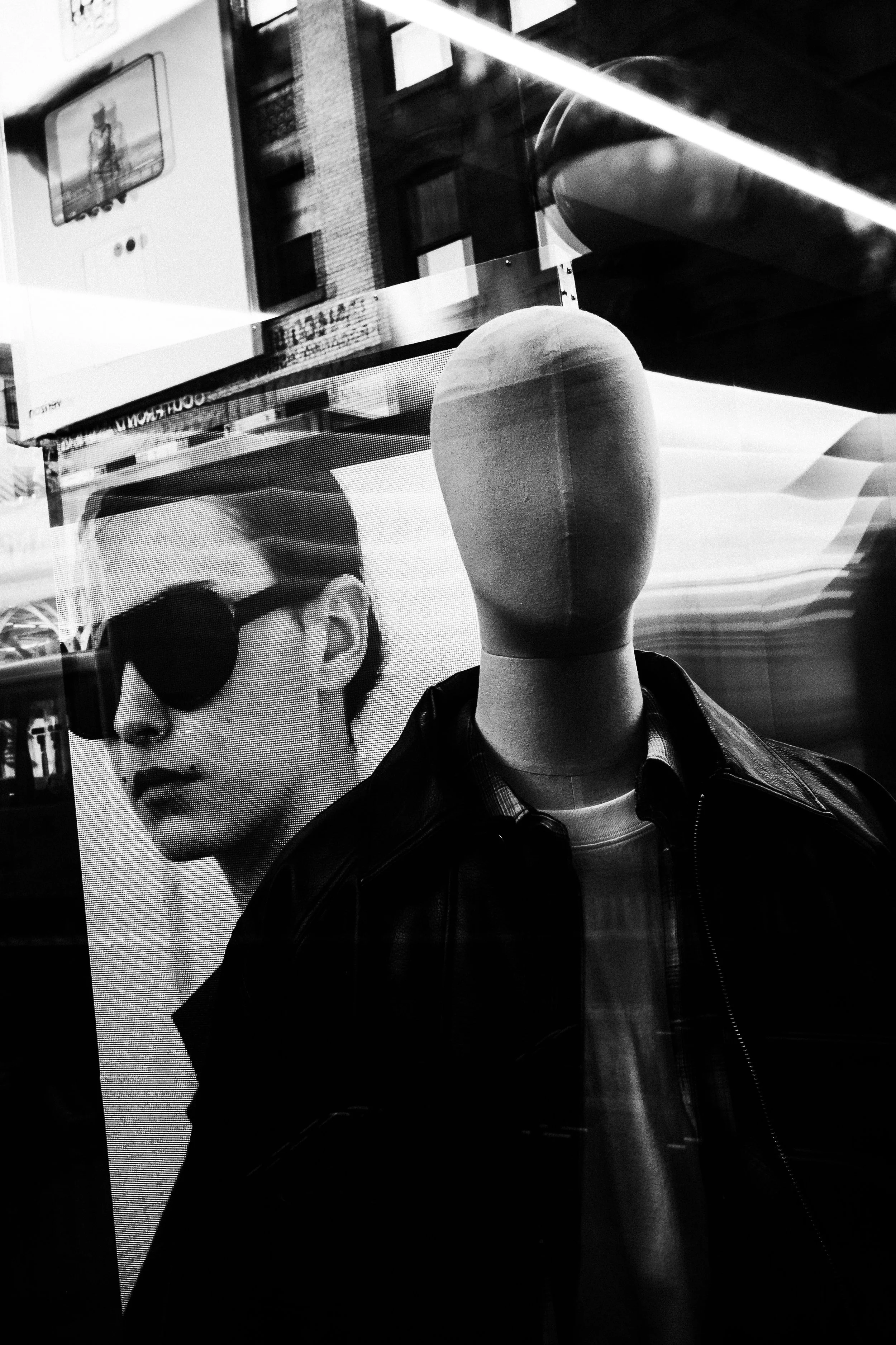 A mannequin with an oval-shaped head wearing a leather jacket and layered clothing, displayed in front of a large digital screen showing a woman in sunglasses.