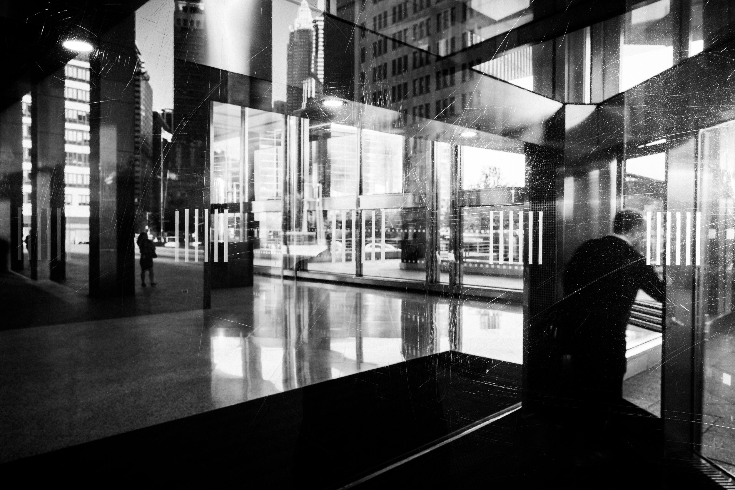 Black and white photo of an urban interior with large glass windows reflecting city buildings. The view includes a person standing outside and the downtown skyline.