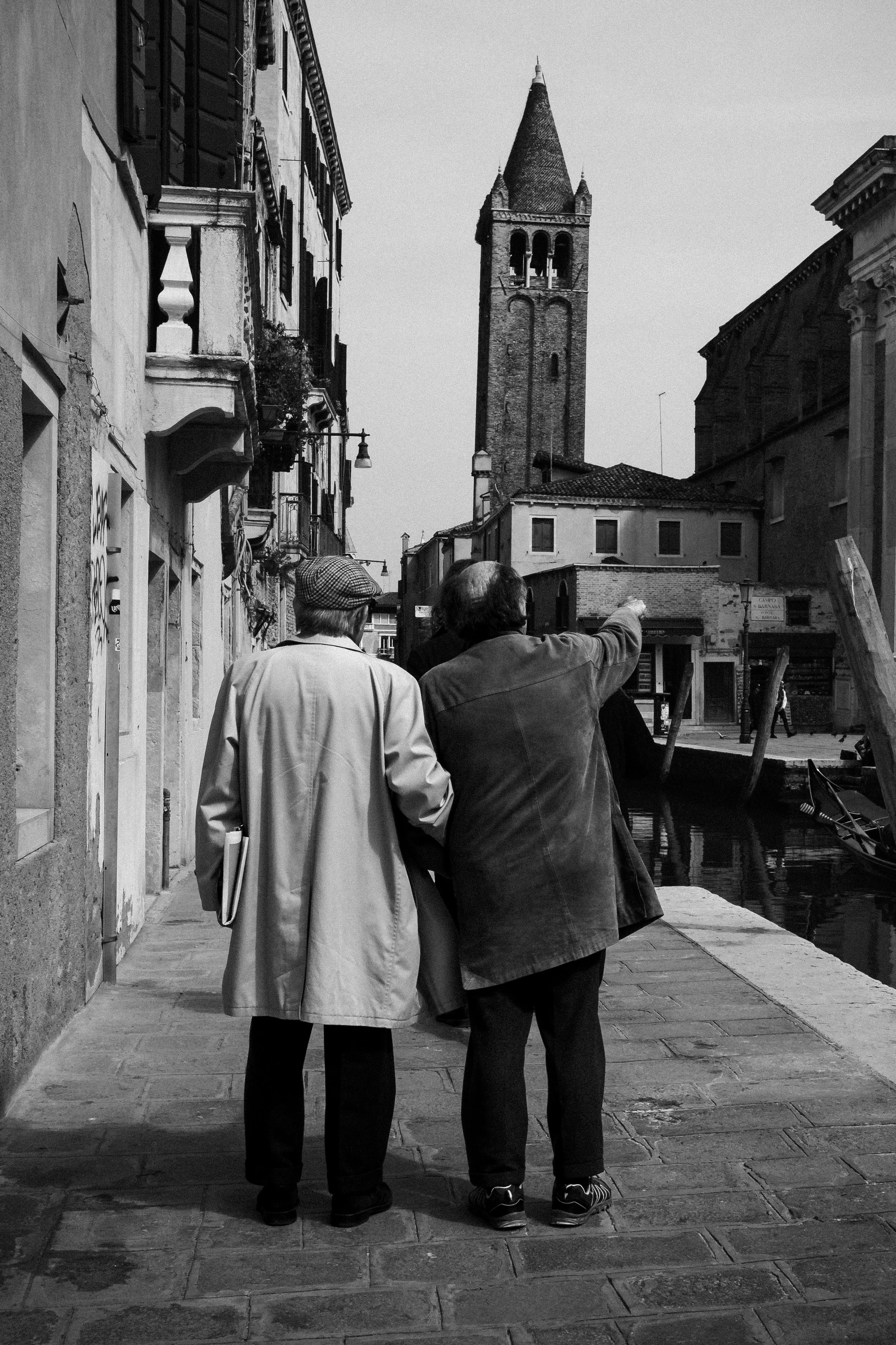 Two people, an older woman and a man, walking along a canal street in Italy, with historic buildings and a church tower in the background, in black and white.