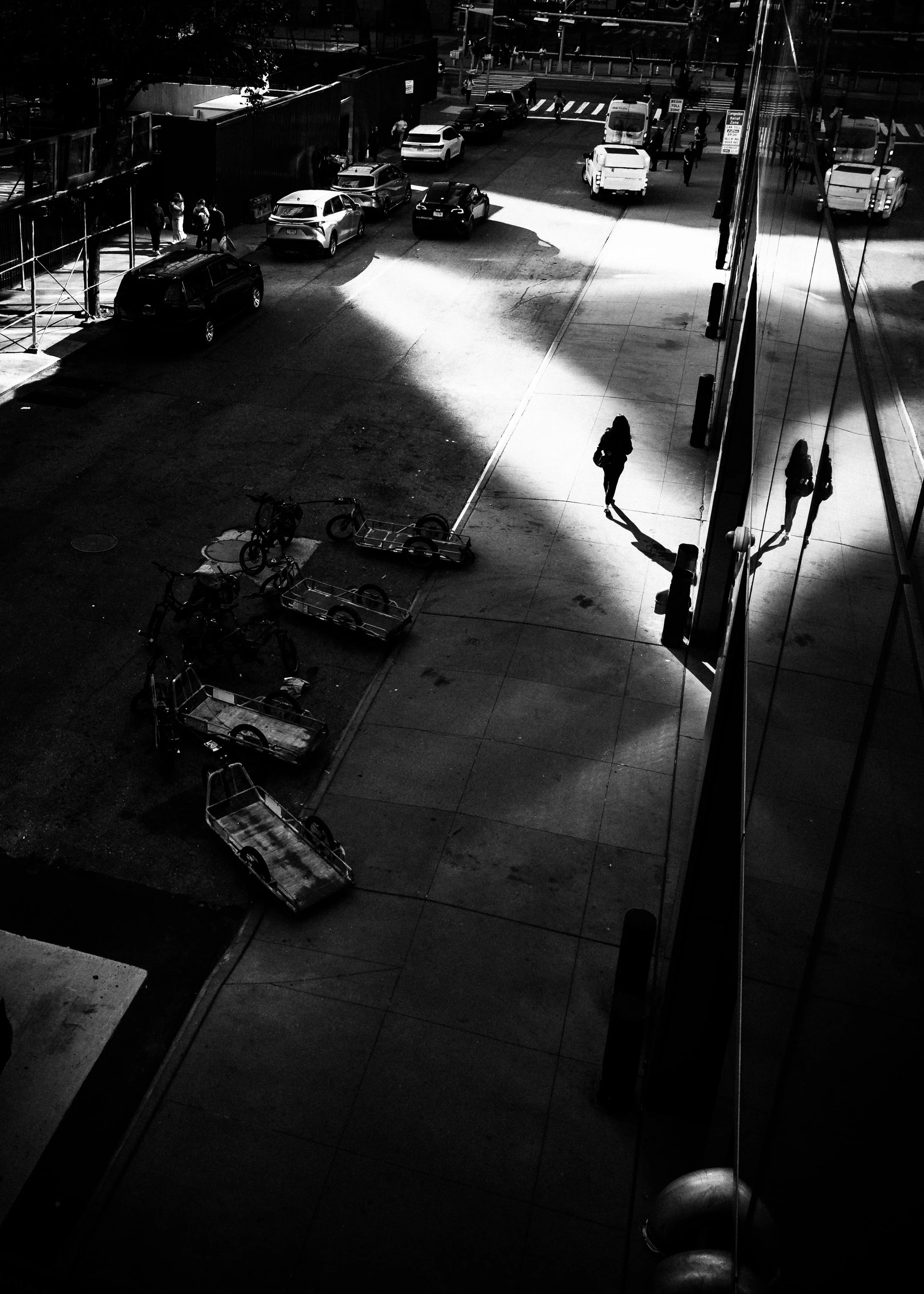 Nighttime street scene with parked cars and pedestrians, divided by bright light and shadow, reflected on a glass building on the right.