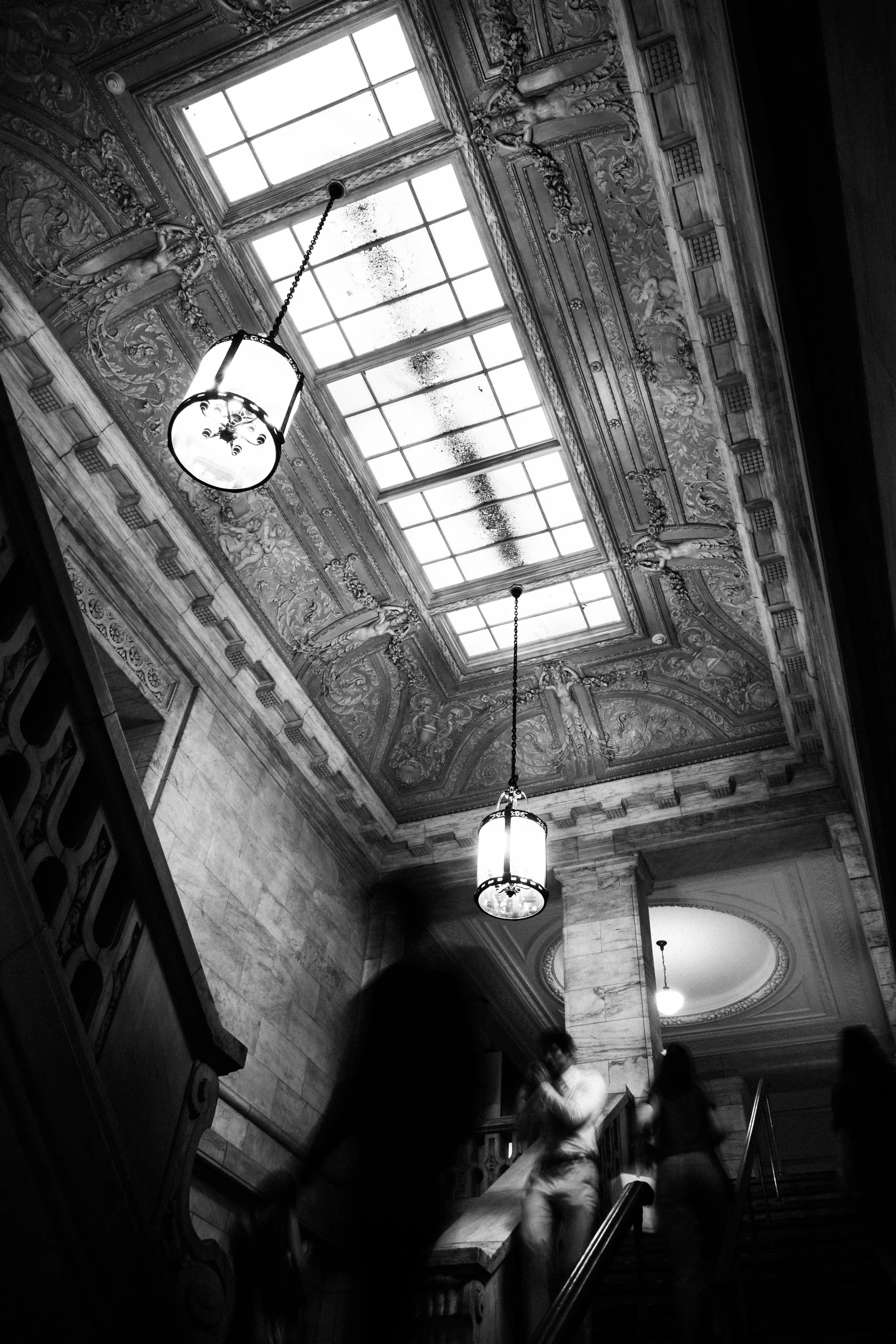 The photo shows a grand, ornately decorated ceiling with a large set of windows allowing natural light to brighten the room. There are decorative elements including sculptures of figures and intricate floral carvings. Three hanging light fixtures are