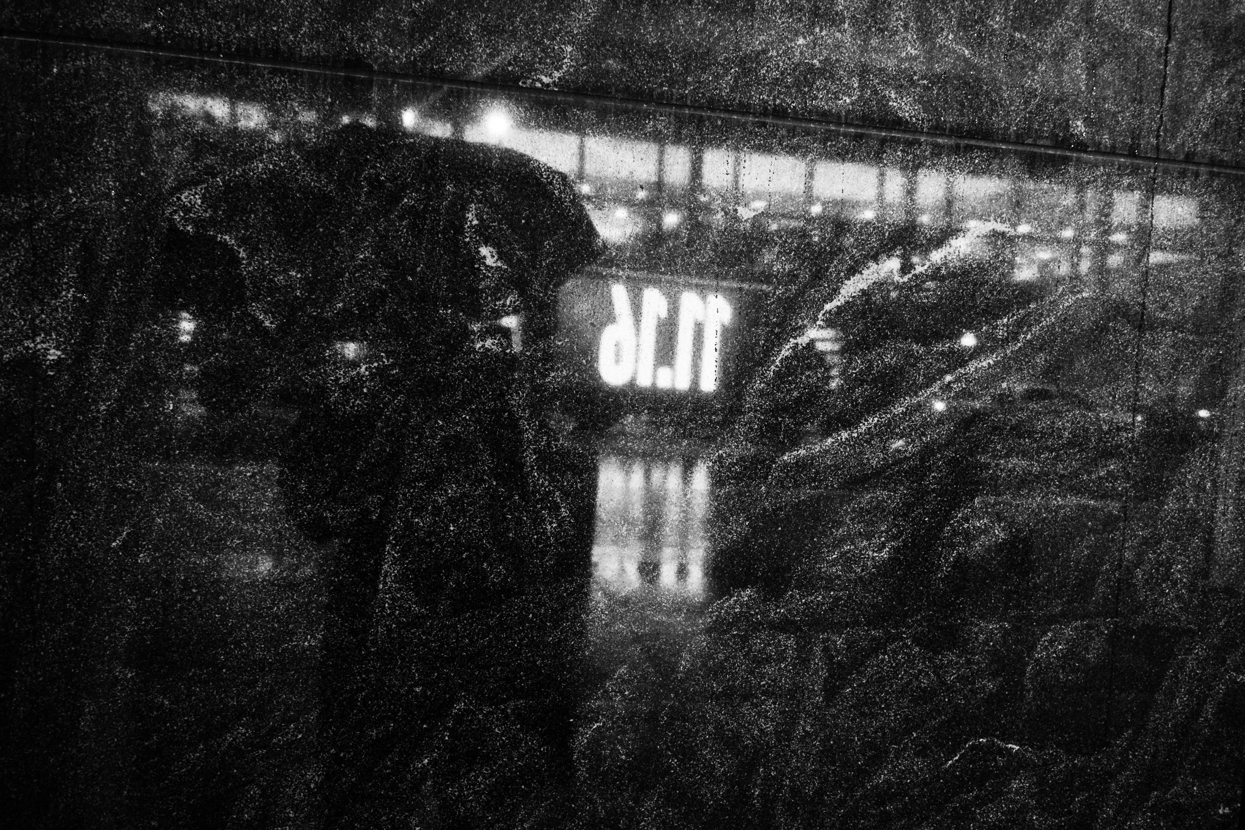 A person wearing a dark coat, standing in the rain behind a rain-covered glass window, with reflections of streetlights and traffic visible.