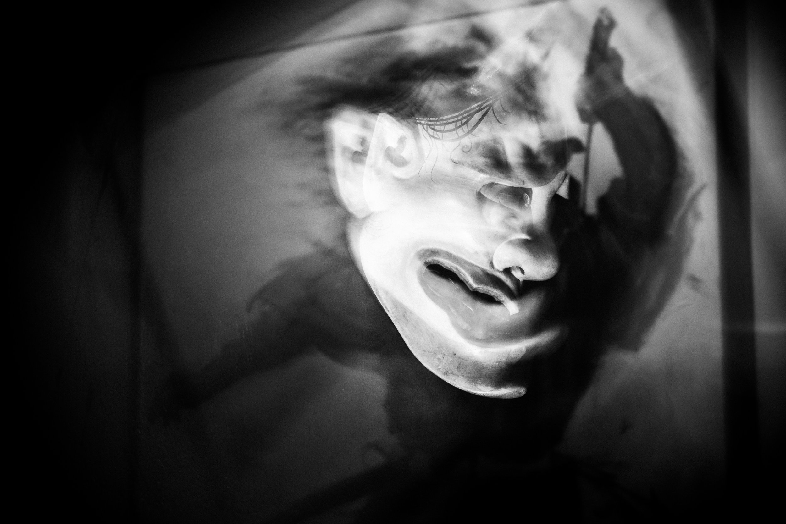A black and white photo of a traditional mask with a serene expression, with flames reflected on its surface.