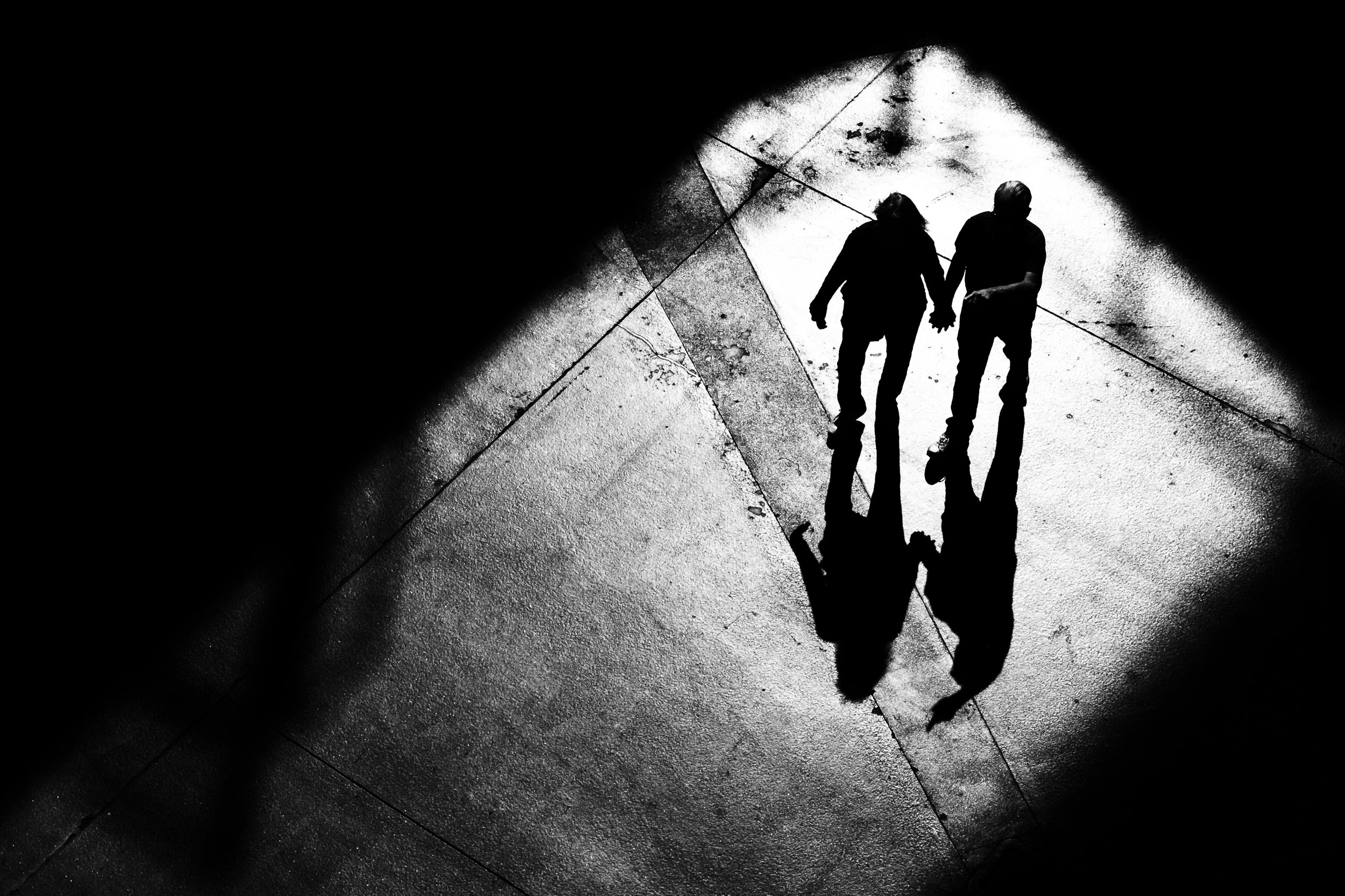 Black and white photo of a couple walking hand in hand on a wet pavement, their reflections visible on the surface.