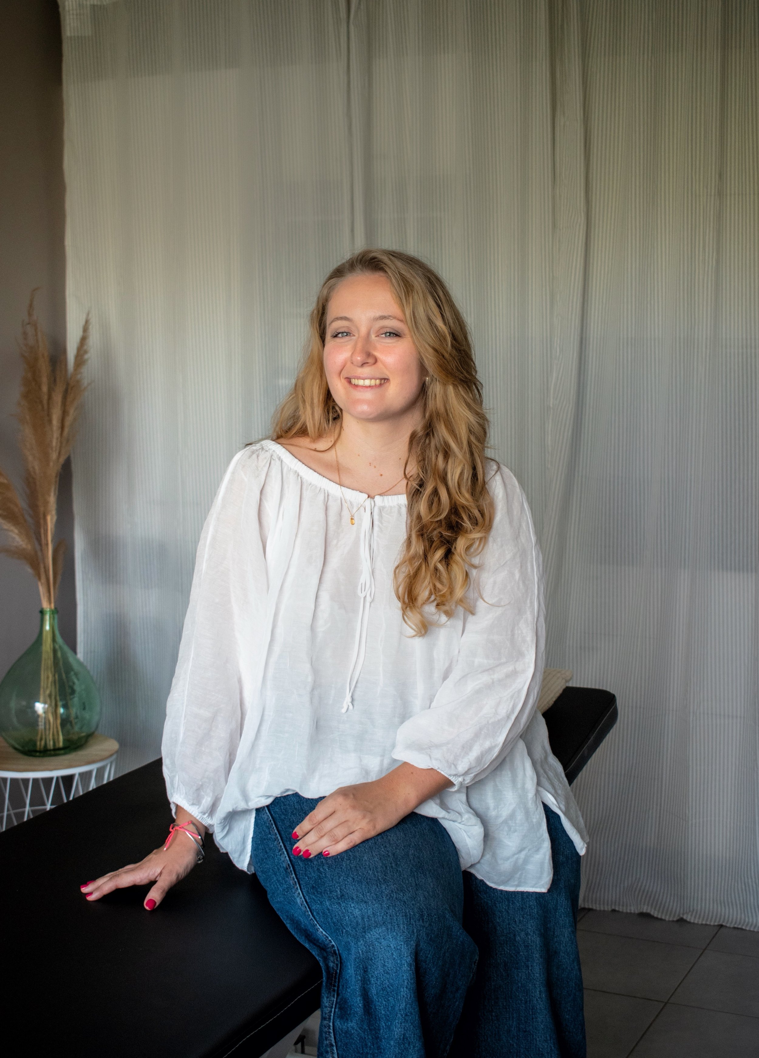Femme souriante aux cheveux blonds, vêtue d’un chemisier blanc et d’un jean, assise dans un intérieur lumineux.