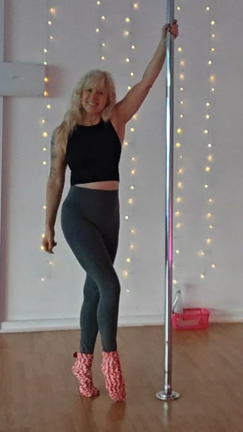 Smiling woman in black crop top, gray leggings, and pink platform boots posing beside a chrome dance pole in a studio with string lights.