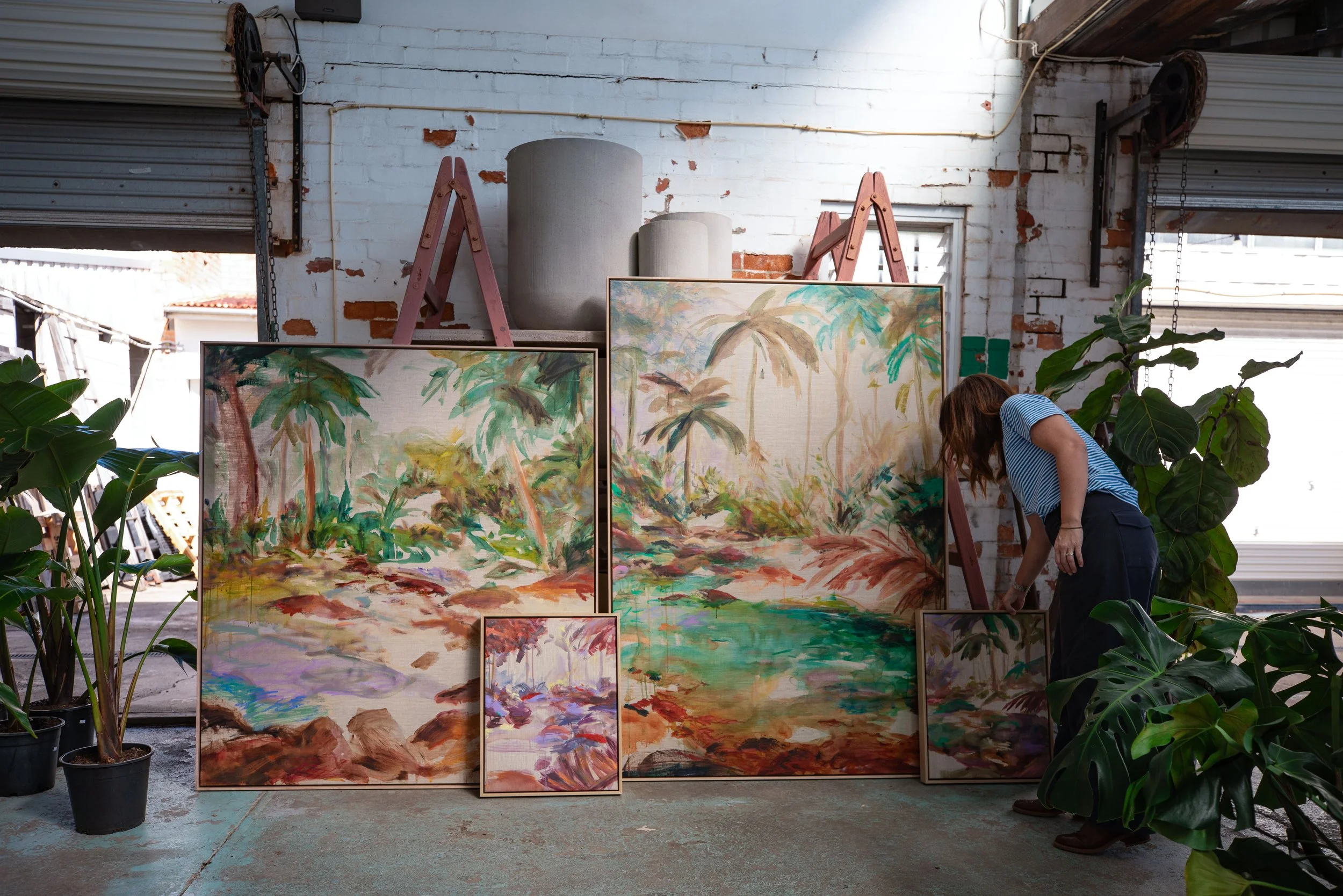 An art gallery featuring landscape paintings with lush greenery and trees, displayed on stands and leaning against a white brick wall. A person with long hair wearing a striped shirt is bending forward to look at the artwork, with large potted plants surrounding the area inside an industrial-style space with open garage doors.