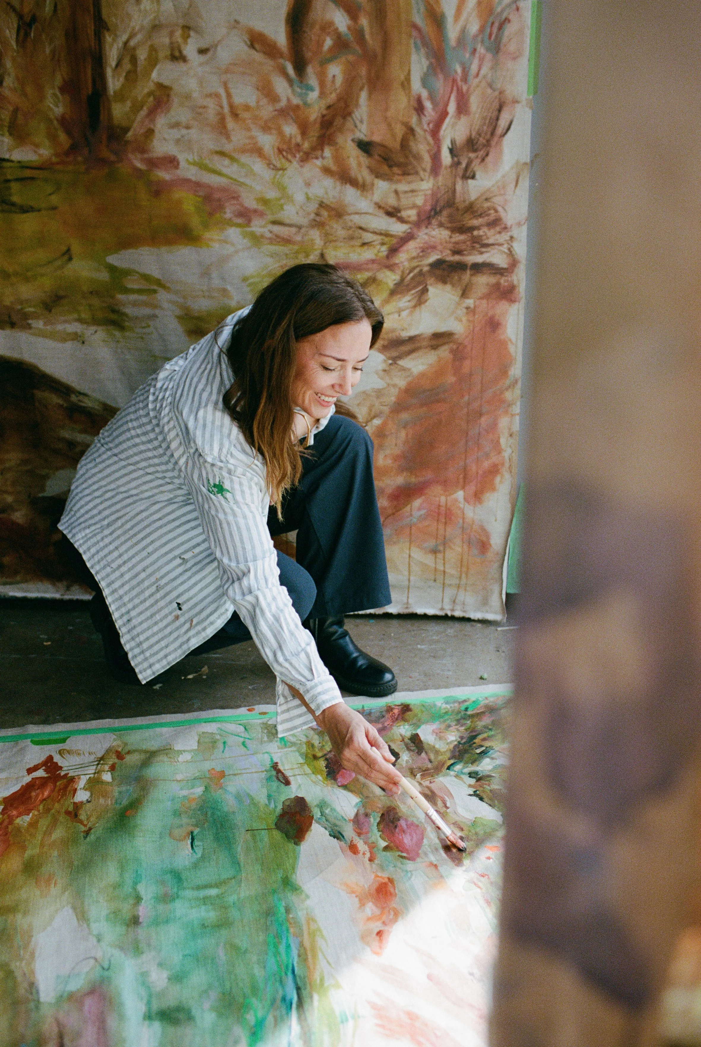 A woman in a striped shirt and dark pants is sitting on the floor, smiling while painting a large watercolor on the floor. The painting features abstract, colorful strokes with probably floral elements. She is holding a paintbrush, and a large, partially finished painting is in the background.