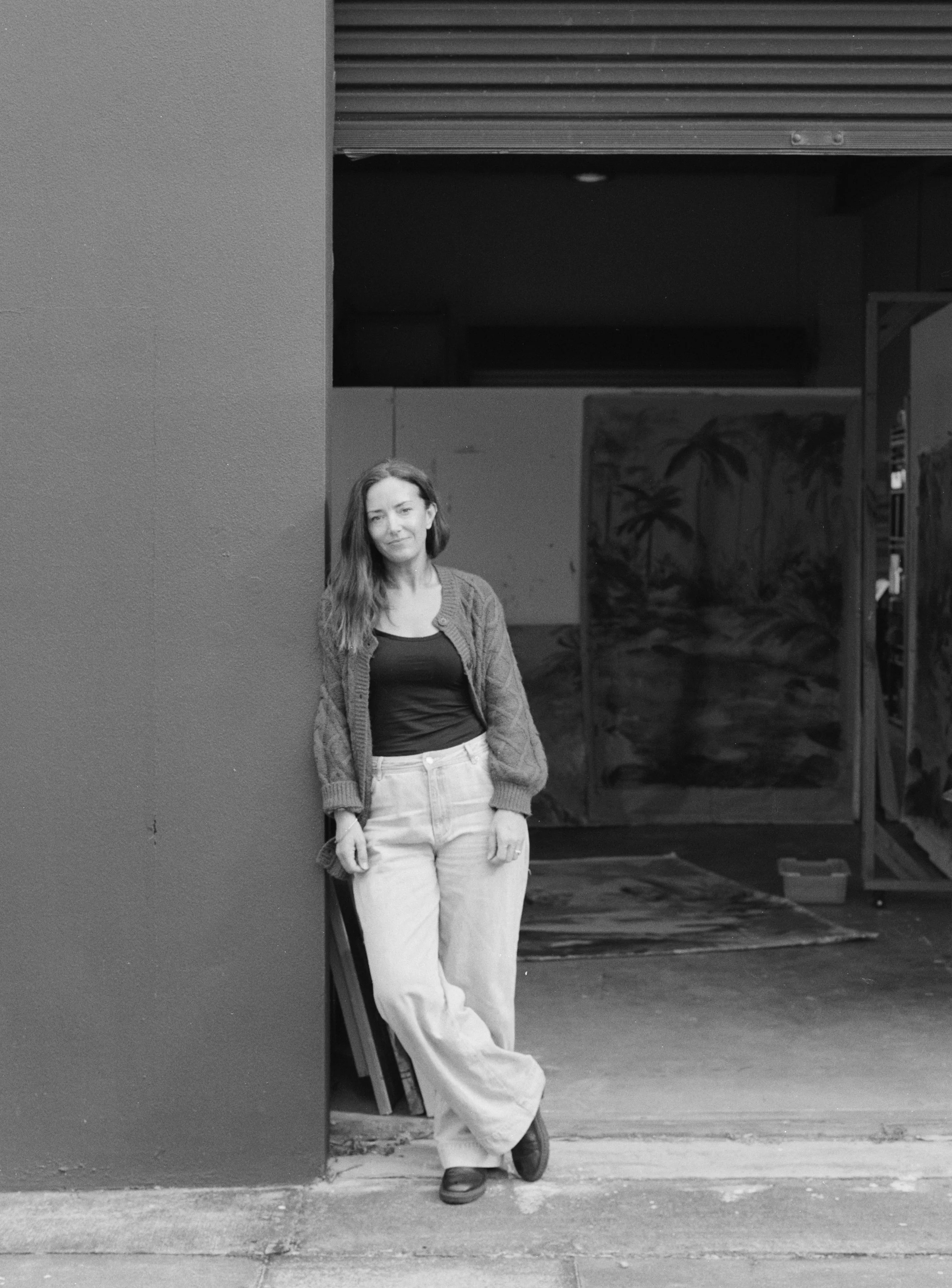 A woman with long hair wearing a sweater, black shirt, white pants, and boots standing with her back against a wall, looking at the camera. She is in front of a construction site with an open garage and some plants painted on the wall inside.