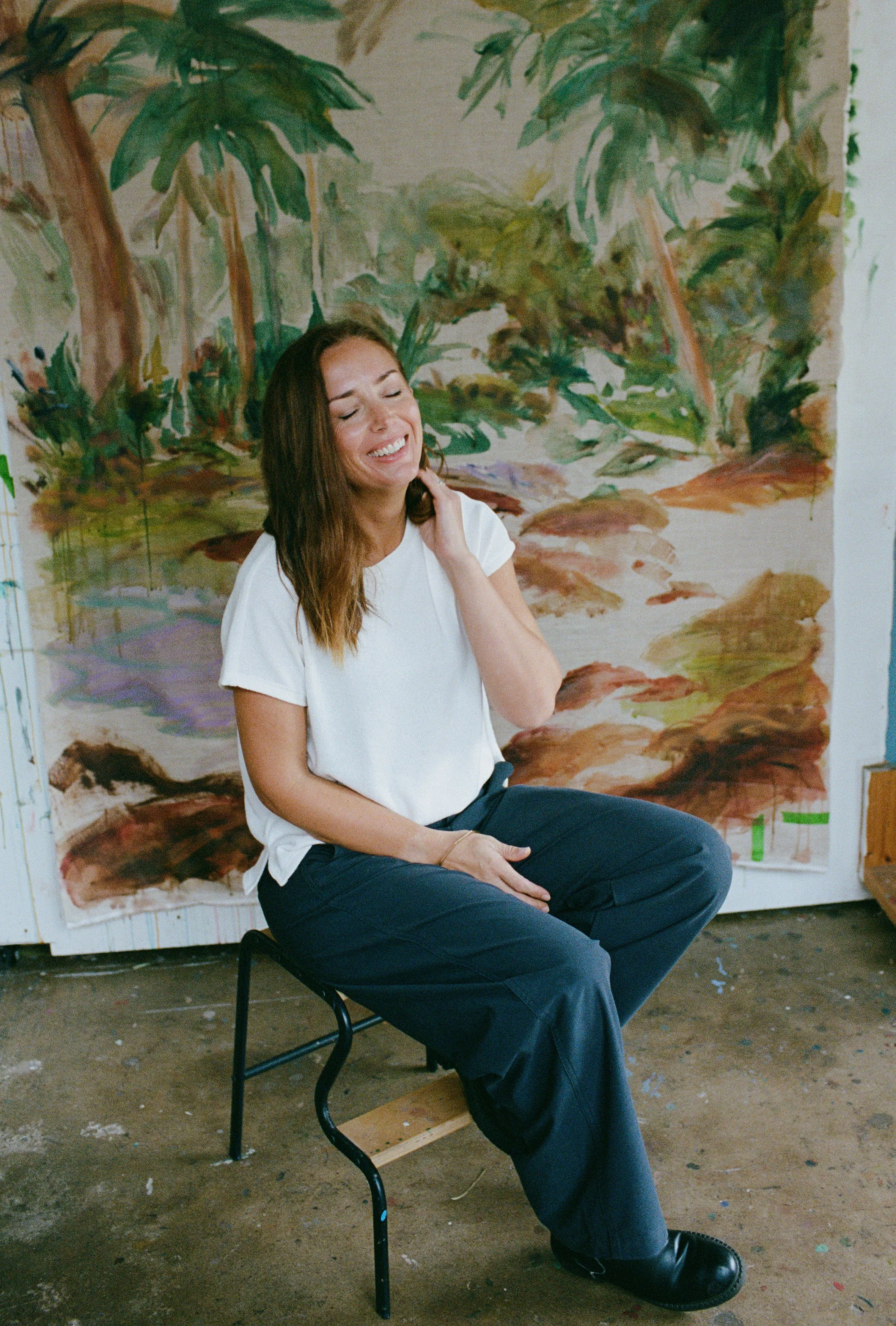 A woman with long brown hair sitting on a chair in front of a large abstract painting or mural with greenery and earthy tones, smiling with eyes closed.