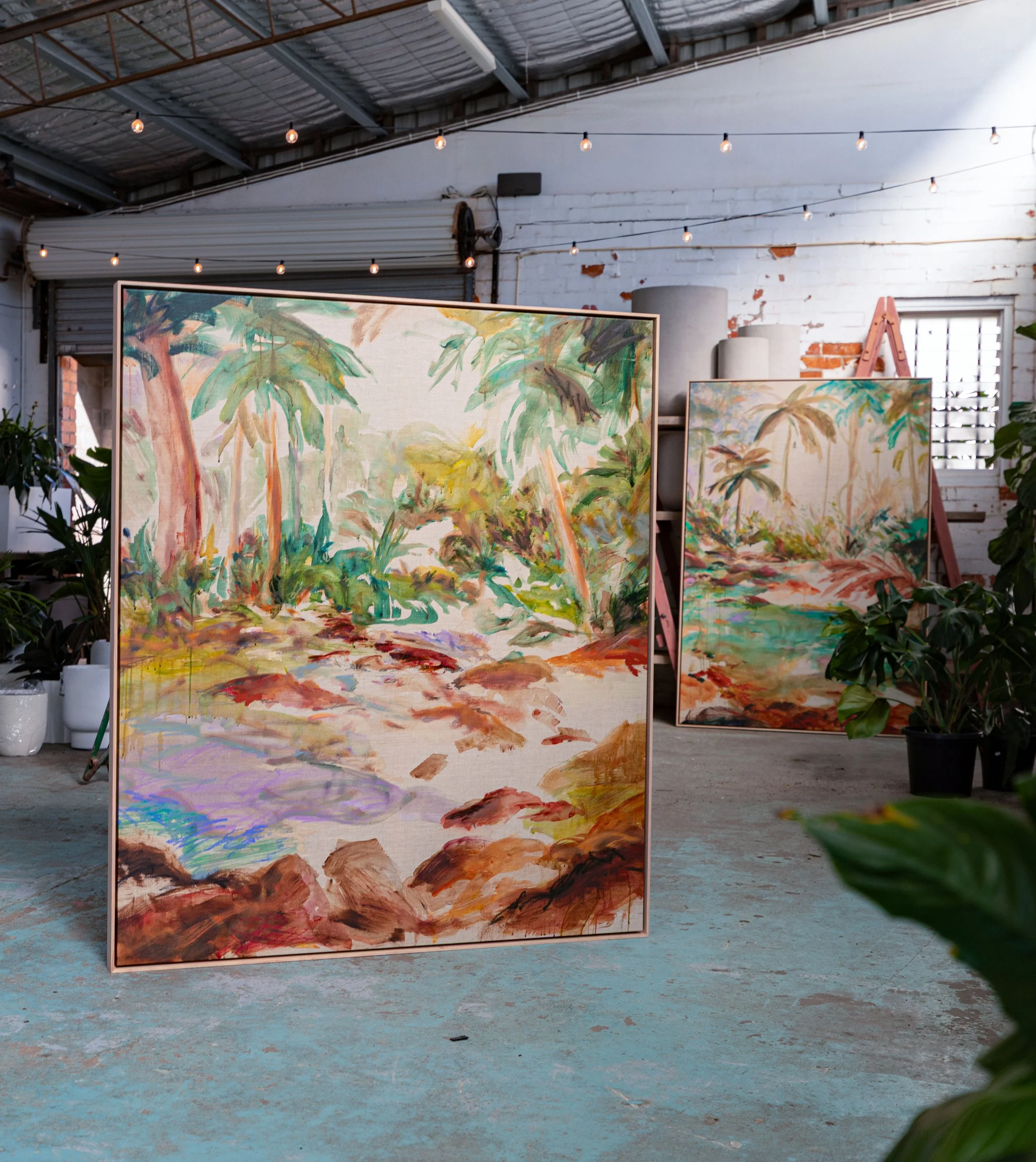 Indoor art gallery displaying colorful landscape paintings on standalone frames, with plants and a brick wall with a window in the background.