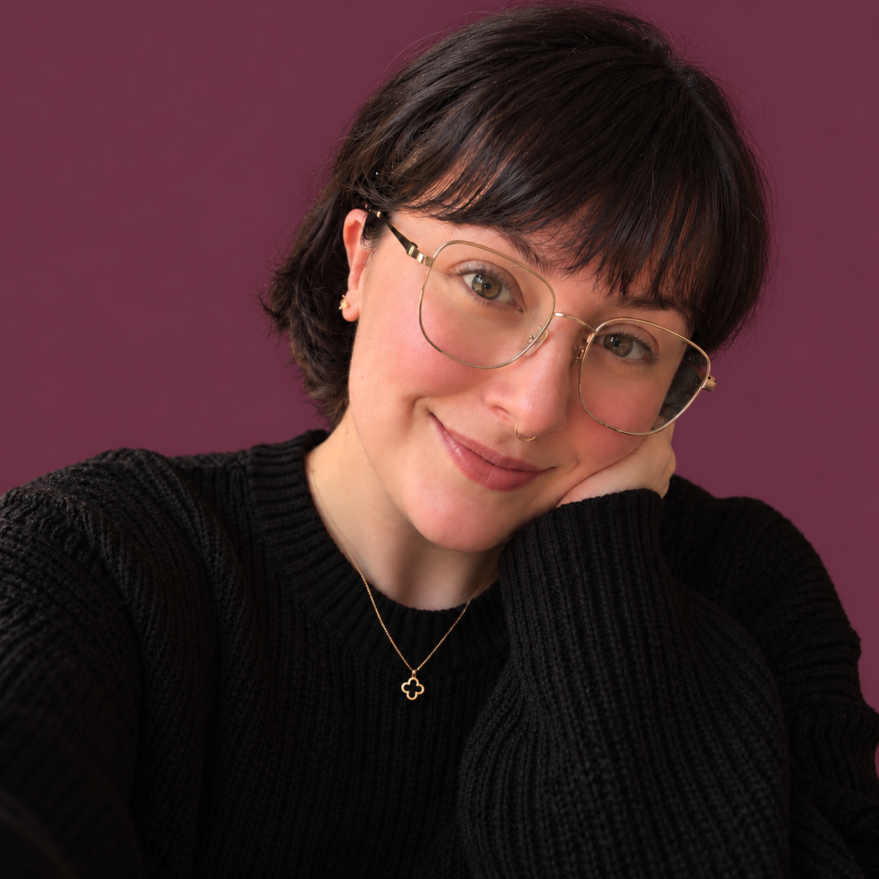A woman with short dark hair, wearing glasses, a black sweater, and a gold cross necklace, resting her face on her hand and smiling at the camera against a purple background.