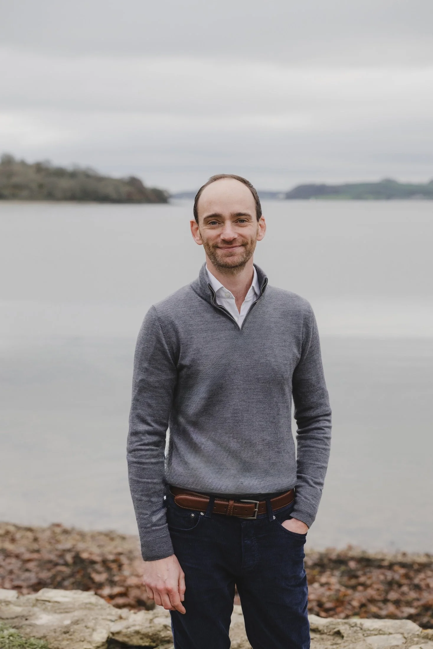 Smiling man in a gray sweater standing by a calm lake with wooded islands.