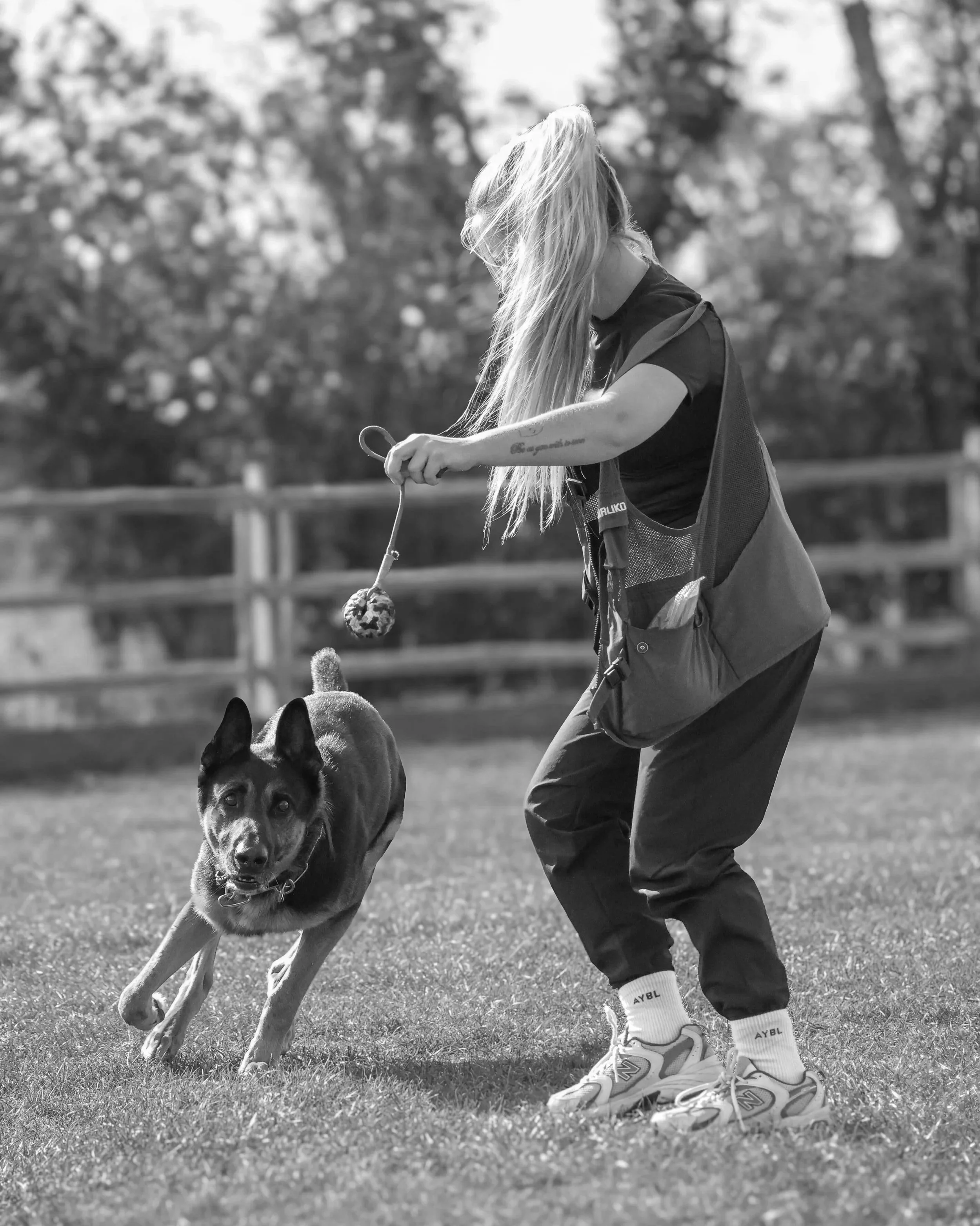 A person with long hair, wearing athletic clothing and sneakers, is playing fetch with a German Shepherd dog outdoors on a grassy field. The person is holding a ball on a string, and the dog is running towards it.