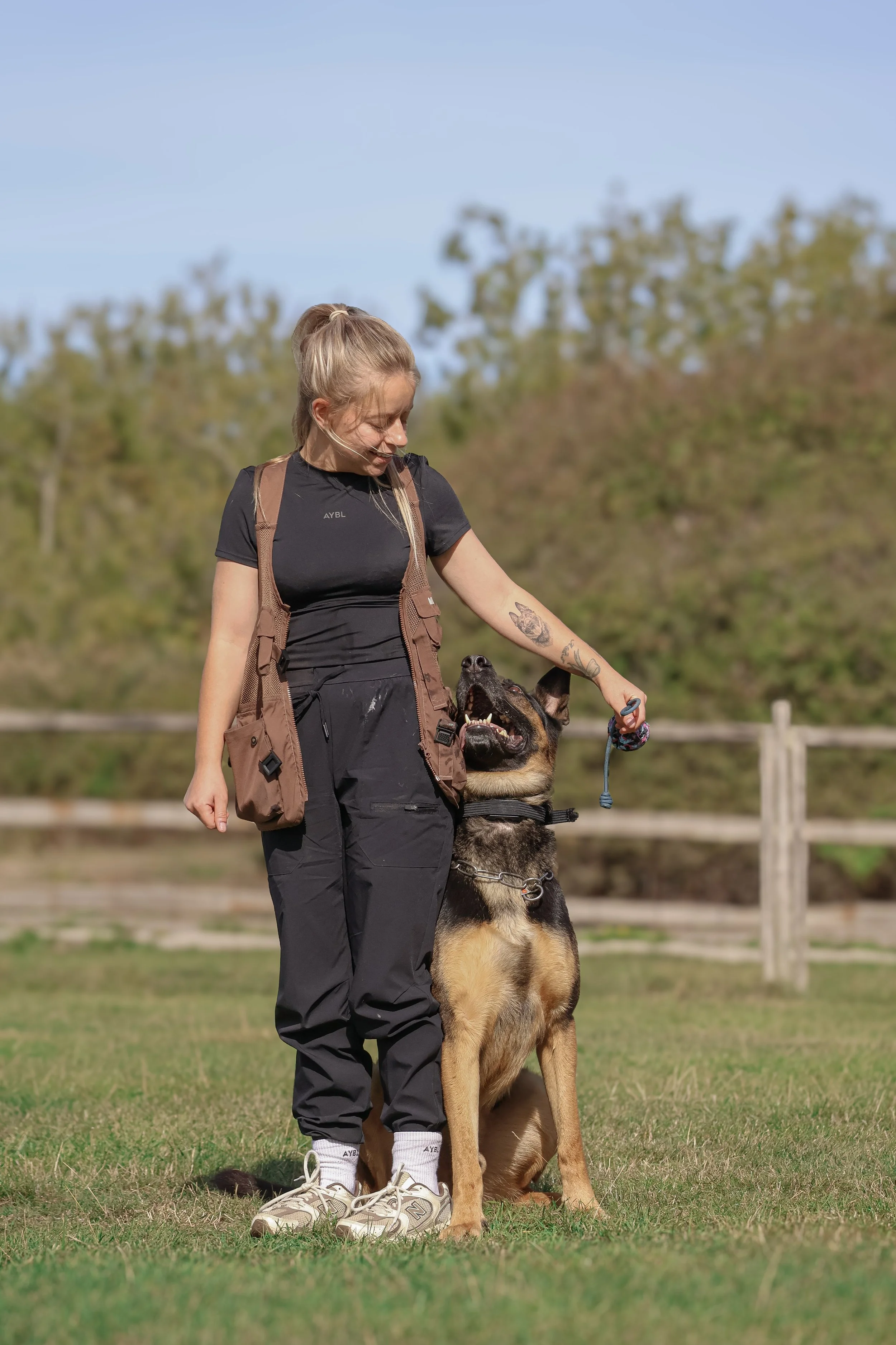 Young woman with a tattoo on her arm standing outdoors with a German Shepherd dog, both appearing happy and engaged.