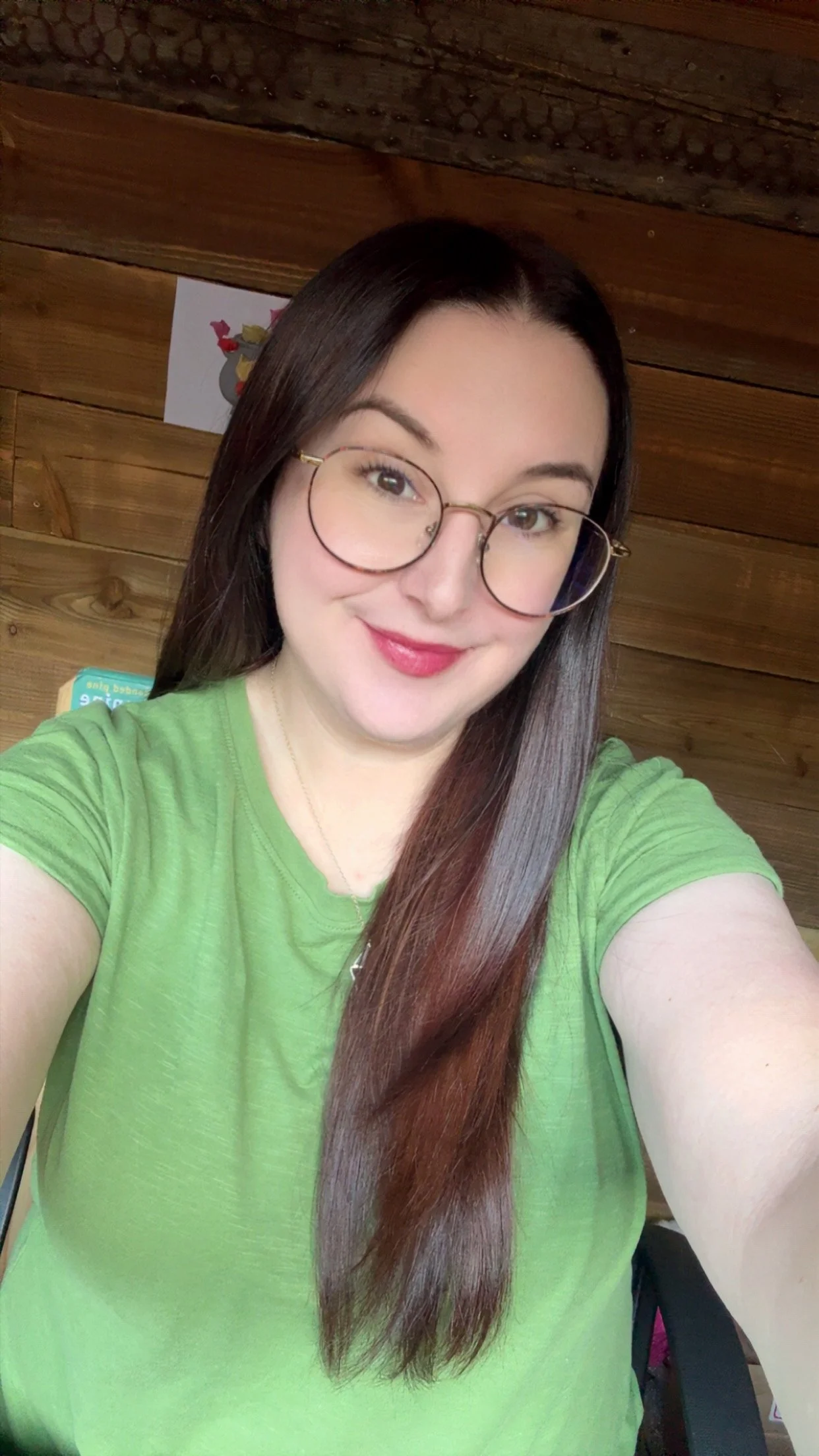 A woman with long brown hair, wearing glasses and a green shirt, taking a selfie indoors in front of a wooden wall.