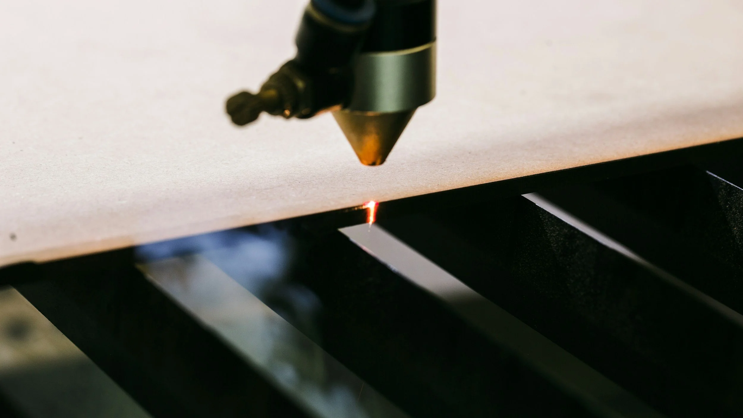 Close-up of a laser cutter or engraver point emitting a red laser beam onto a piece of material, with focus on the laser tip and the illuminated spot on the material surface.