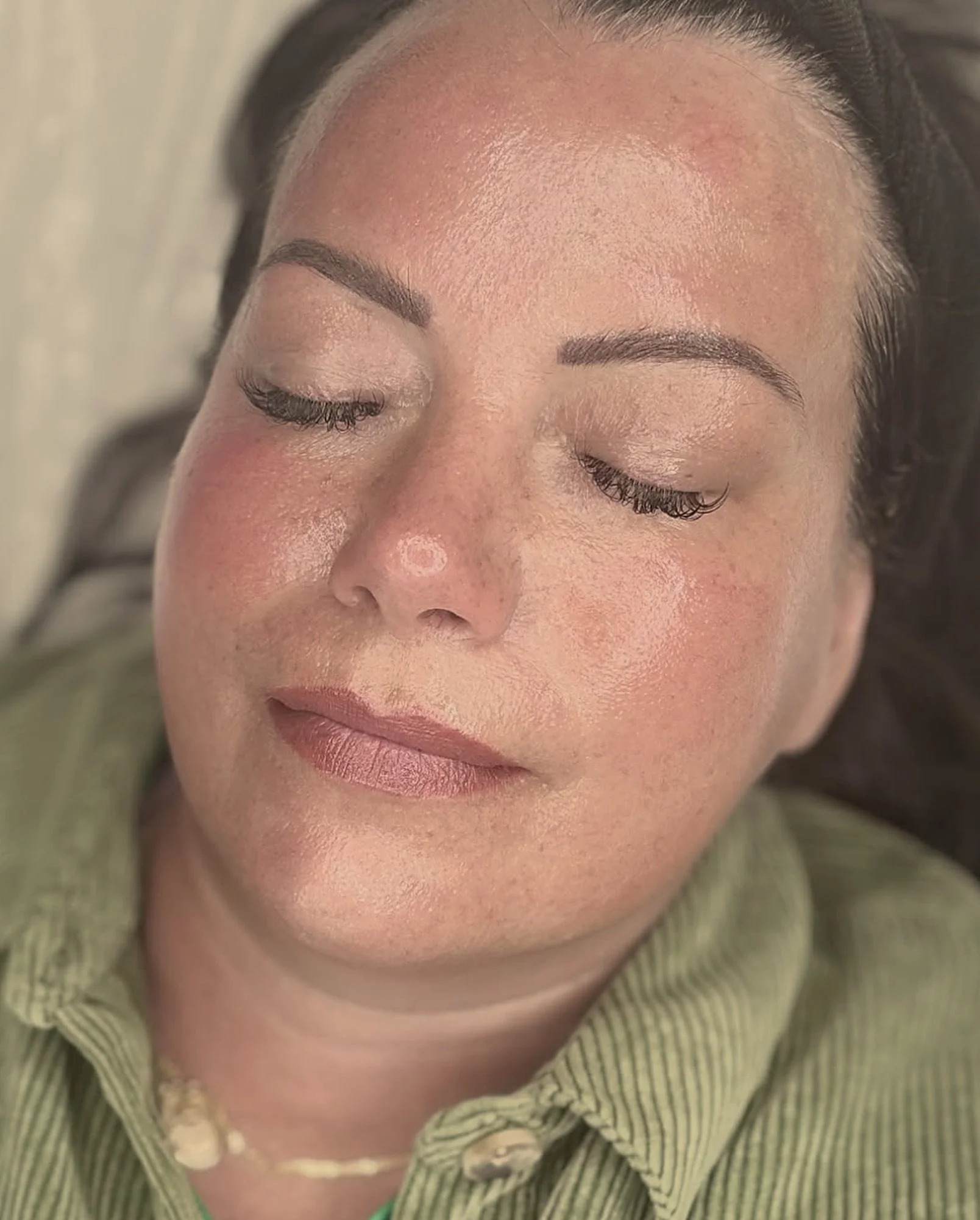 Close-up of a woman's face with her eyes closed, showing shiny skin, well-groomed eyebrows, and curled eyelashes. She is wearing a green striped shirt and a beige scarf.