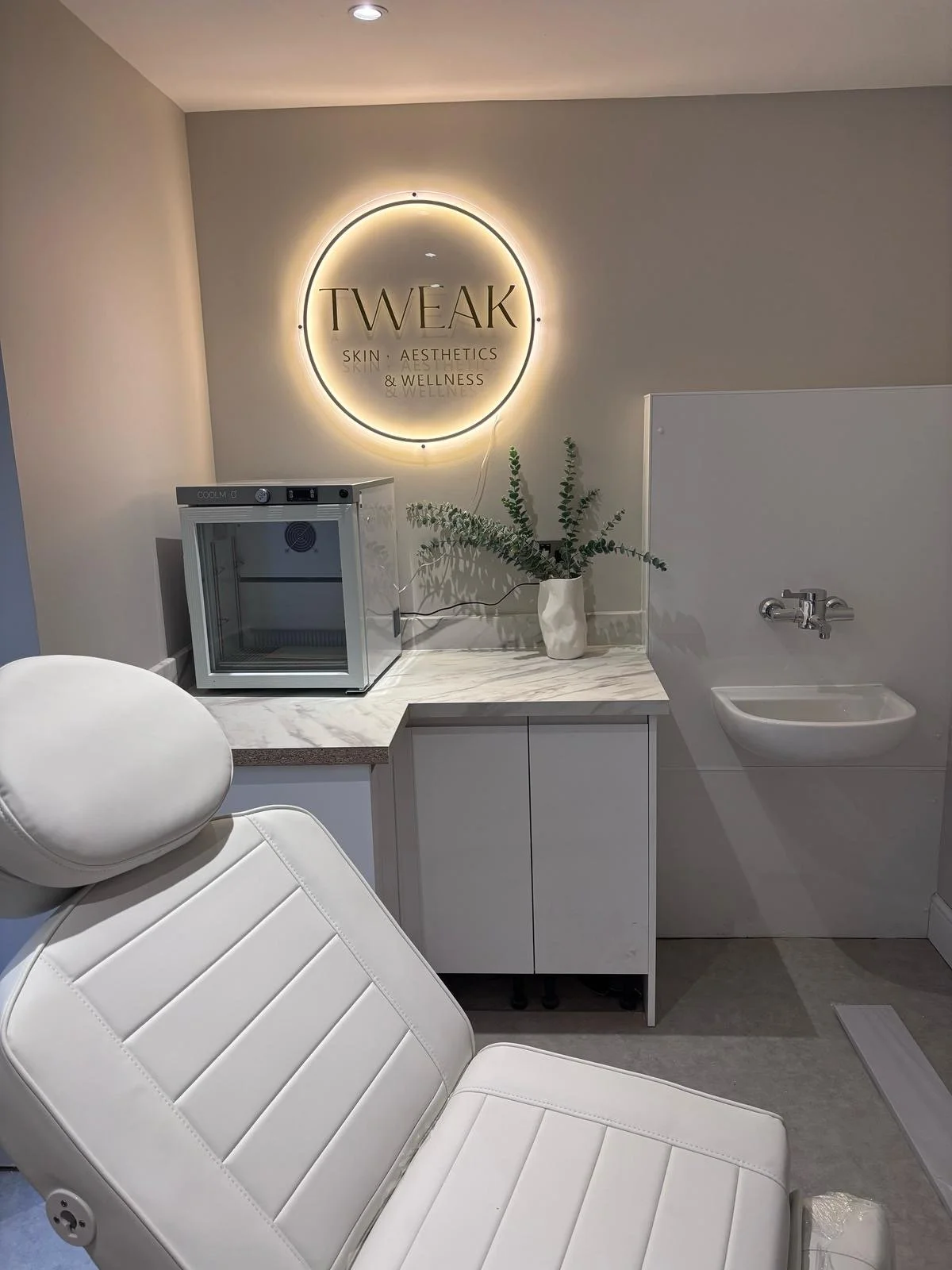 A modern spa or aesthetic clinic interior with a comfortable white treatment chair, a small fridge, a potted plant, and a wall sign that reads 'TWEAK Skin, Aesthetics, & Wellness' illuminated with backlighting.